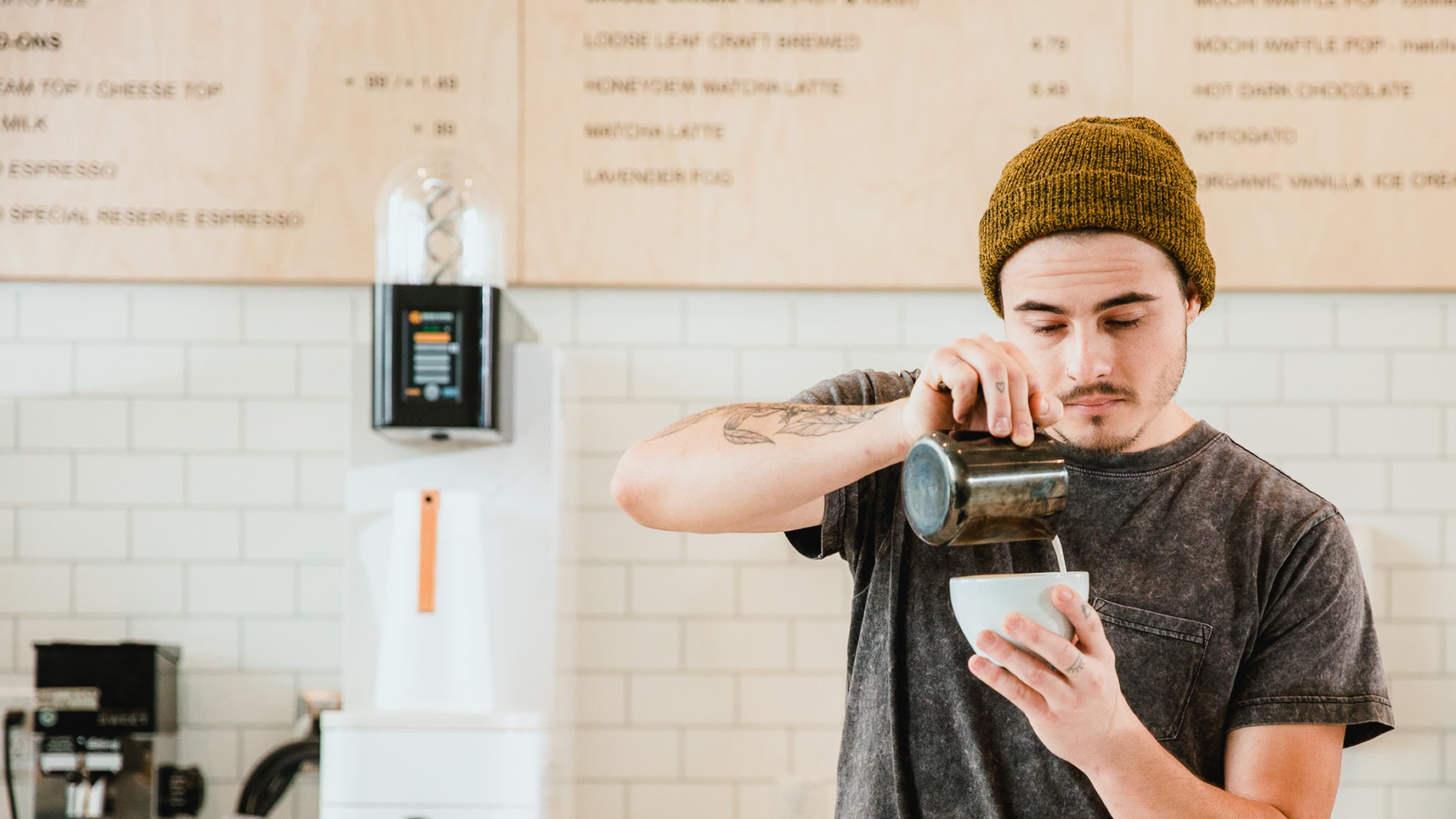 Barista with tattoos and a mustard beanie pours milk into a cup at a coffee shop with a light wood and brick interior.