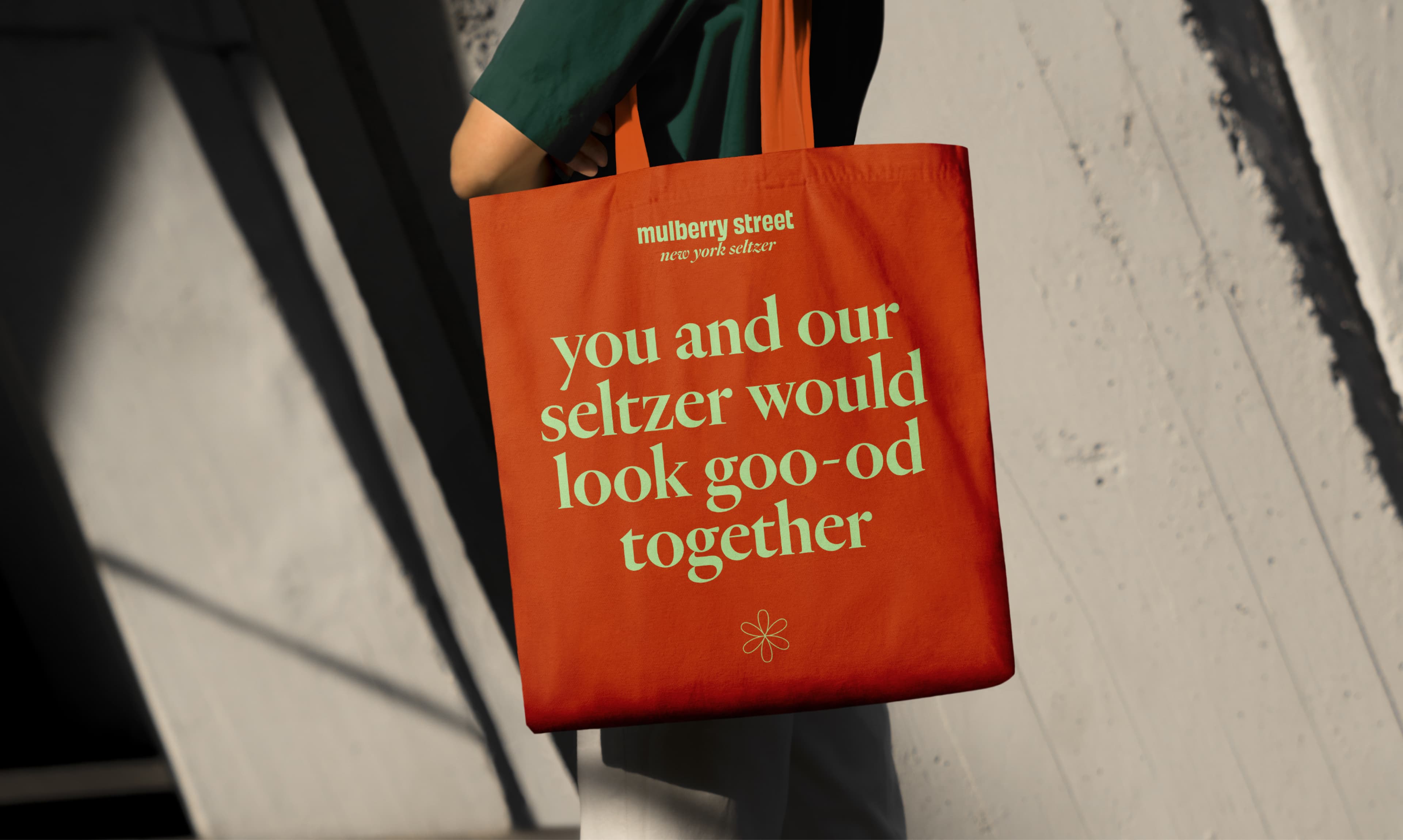Person holding a red tote bag branded "Mulberry Street New York Seltzer" with the tagline "you and our seltzer would look goo-od together" in light green text, against a shadowed wall.