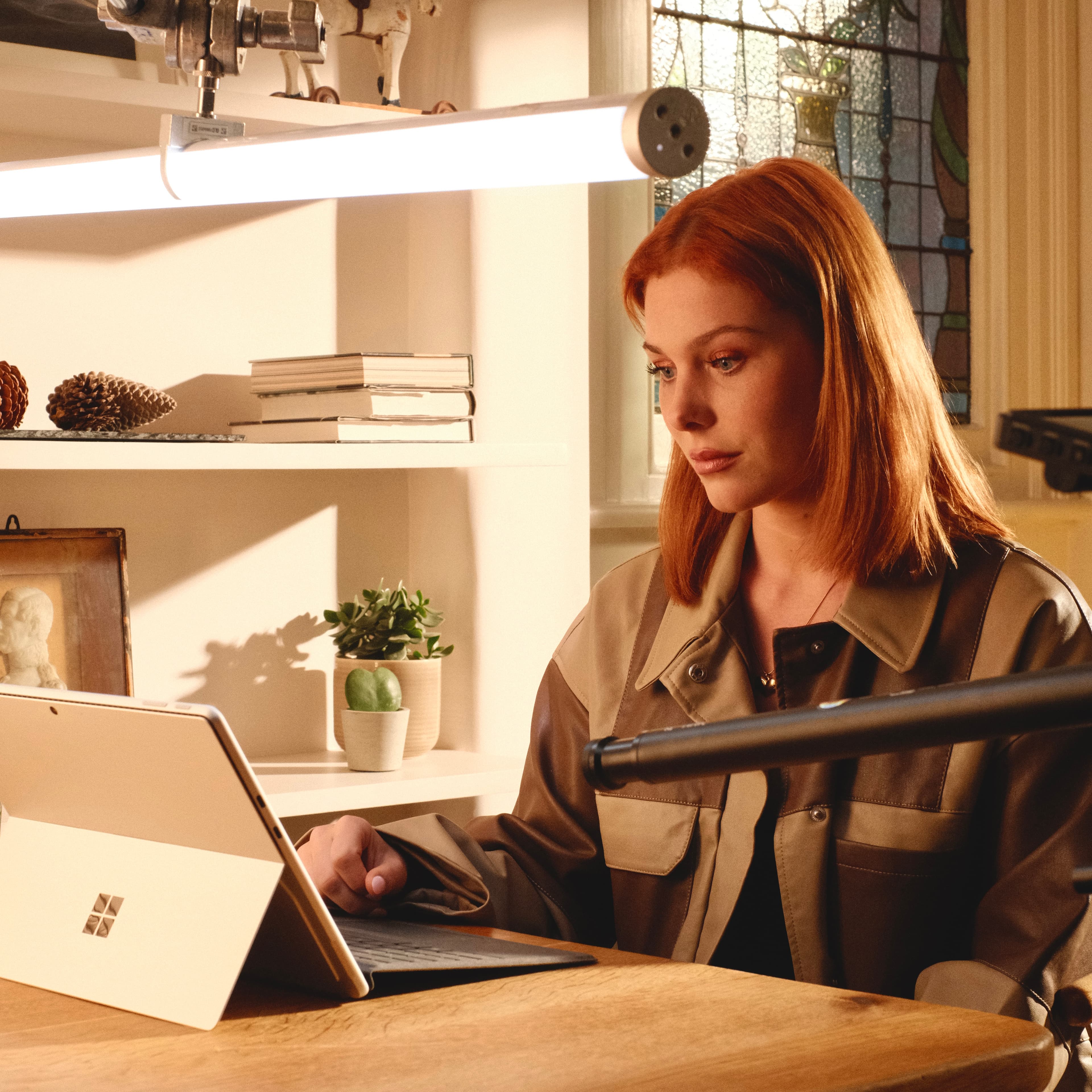 Woman with red hair using a Microsoft Surface laptop at a wooden desk in a warmly lit room with plants and books on shelves.