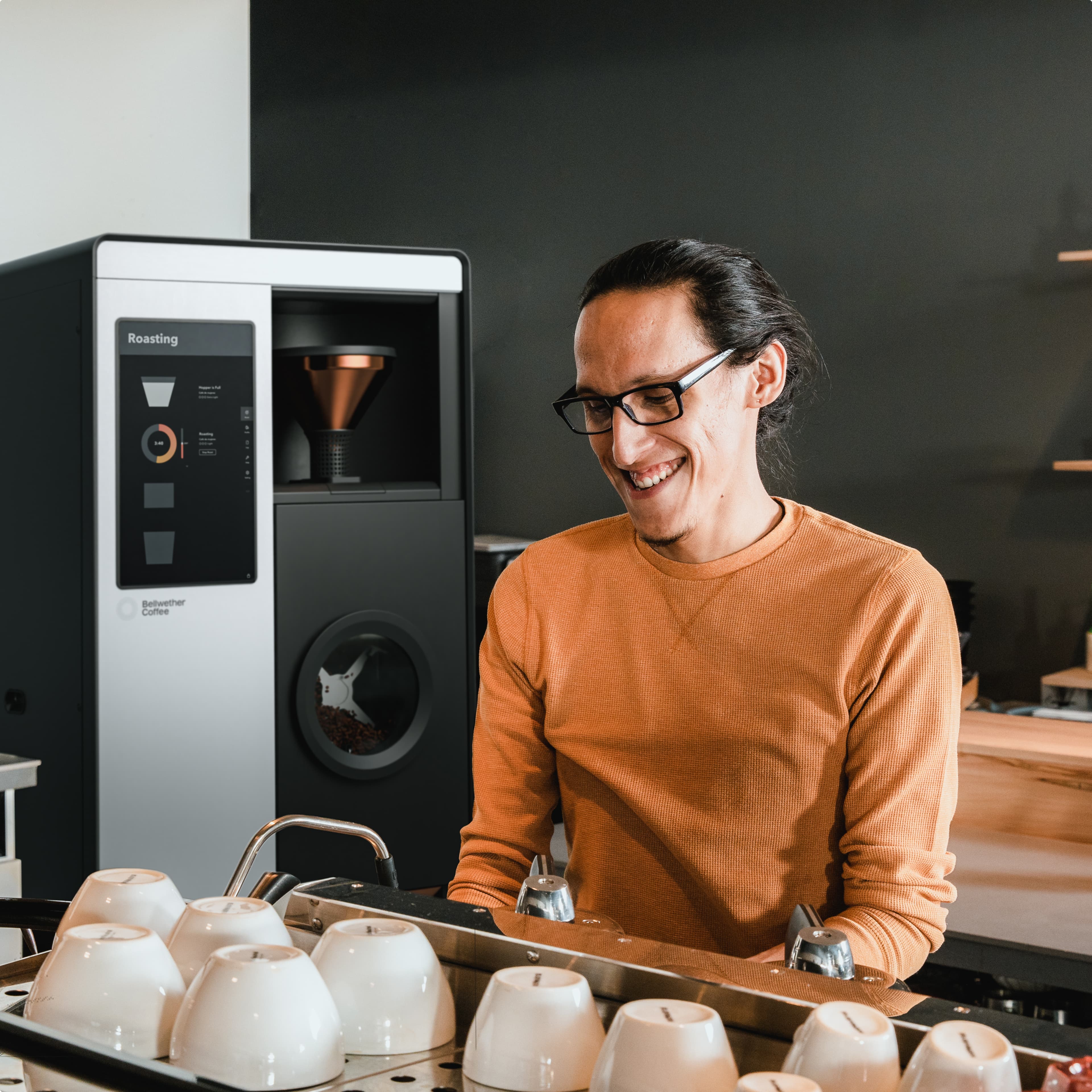 Person with glasses in an orange sweater smiles in front of a coffee roaster with white coffee cups lined up on a tray.