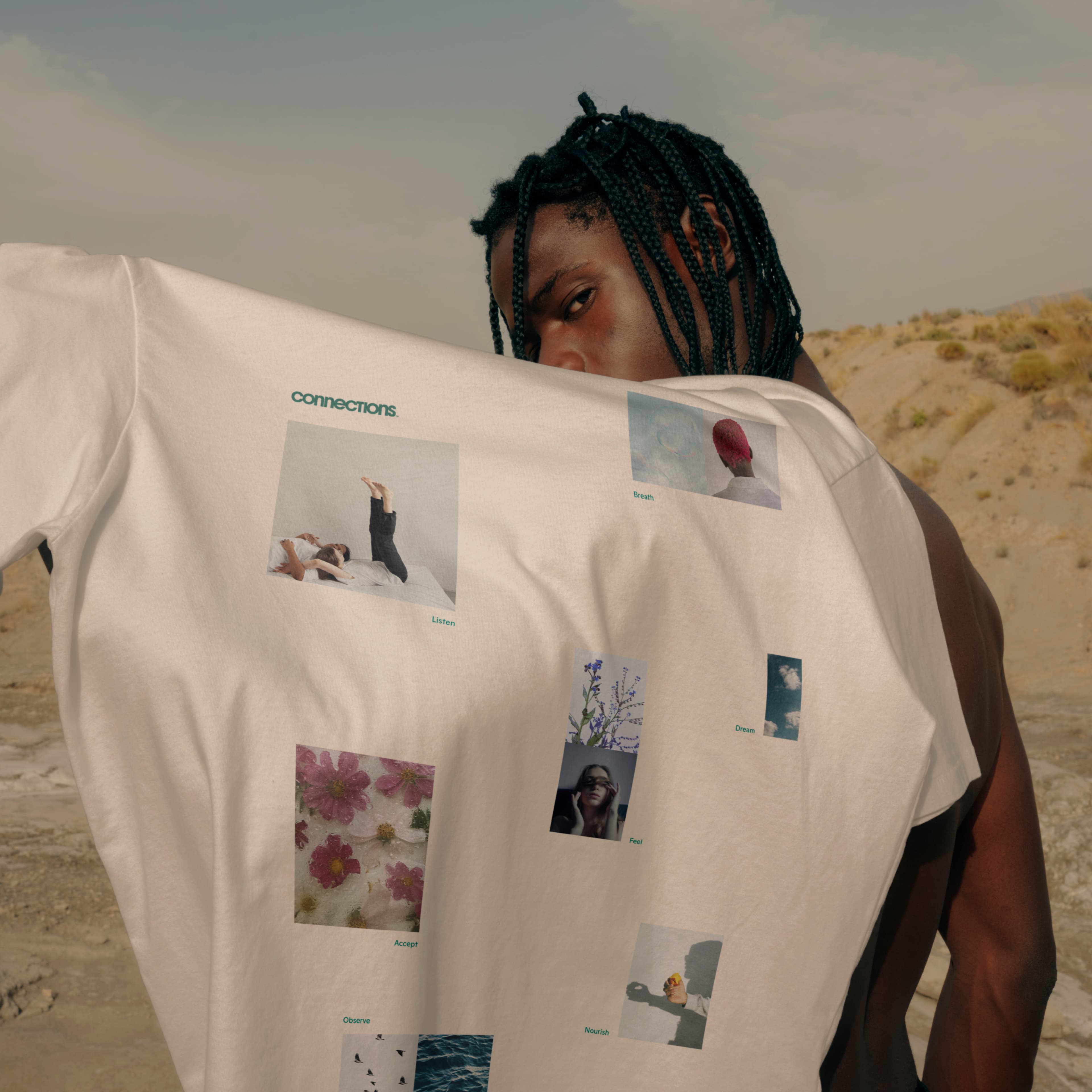 Person holding up the back of a white "Connections" T-shirt printed with artistic photo collages labeled Breathe, Listen, Dream, Feel, Accept, Observe, and Nourish, photographed in a desert landscape.