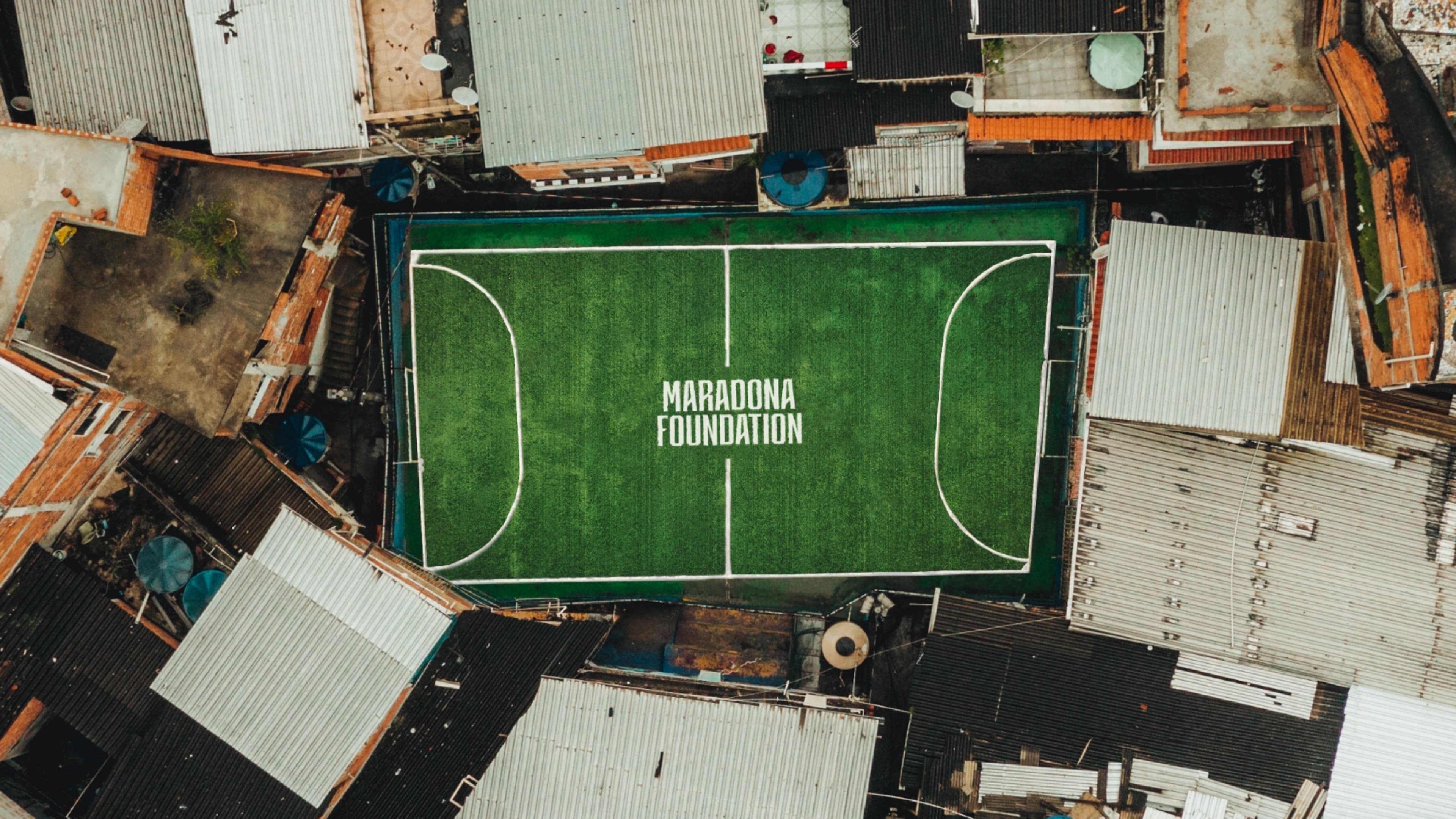 Aerial view of a small soccer field labeled 'Maradona Foundation' surrounded by urban rooftops with corrugated metal and brick buildings.