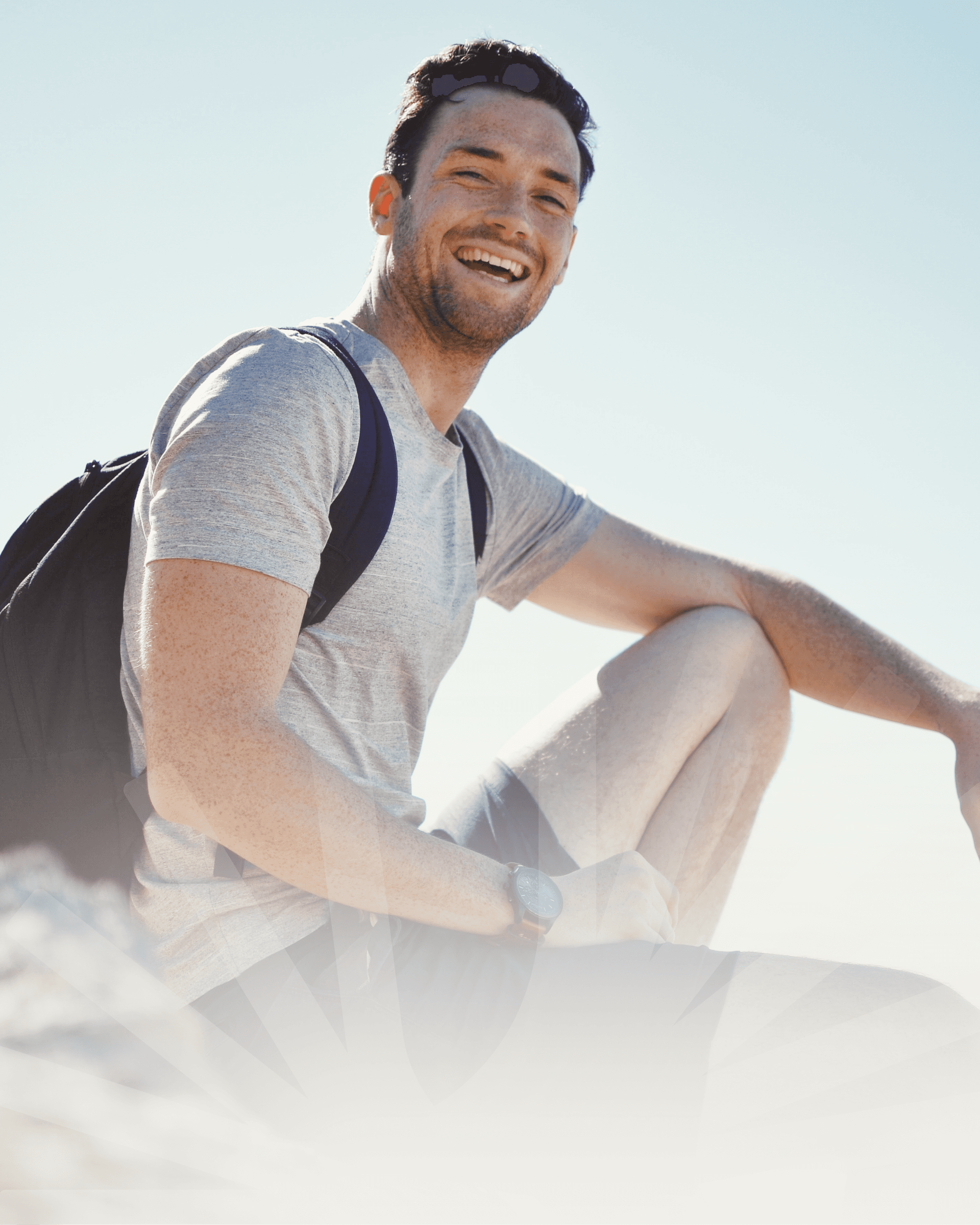 Man with short hair smiling in gray t-shirt and black backpack, sitting on a rock under a clear blue sky on a sunny day.