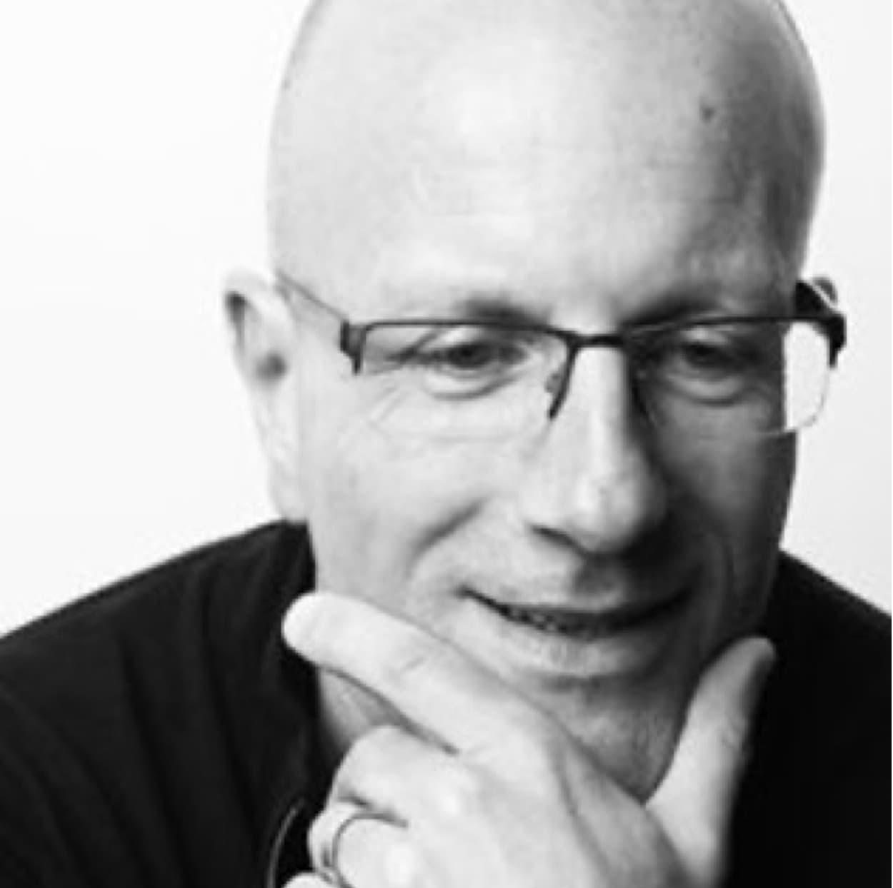 A man in glasses and a black shirt smiles as he is holding his chin.