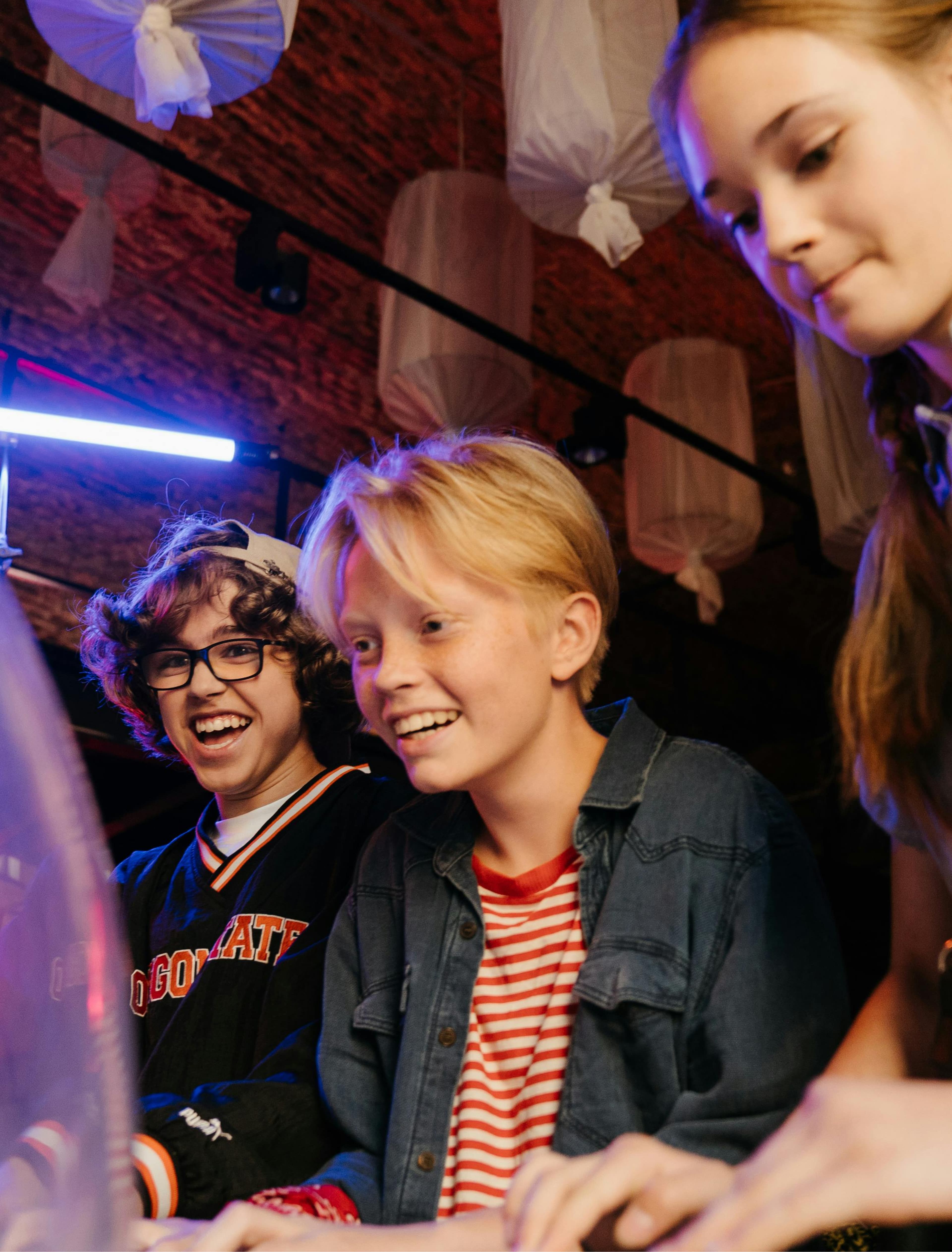 Three kids smiling and playing arcade games with enthusiasm under a ceiling with white fabric decorations and colorful lighting.
