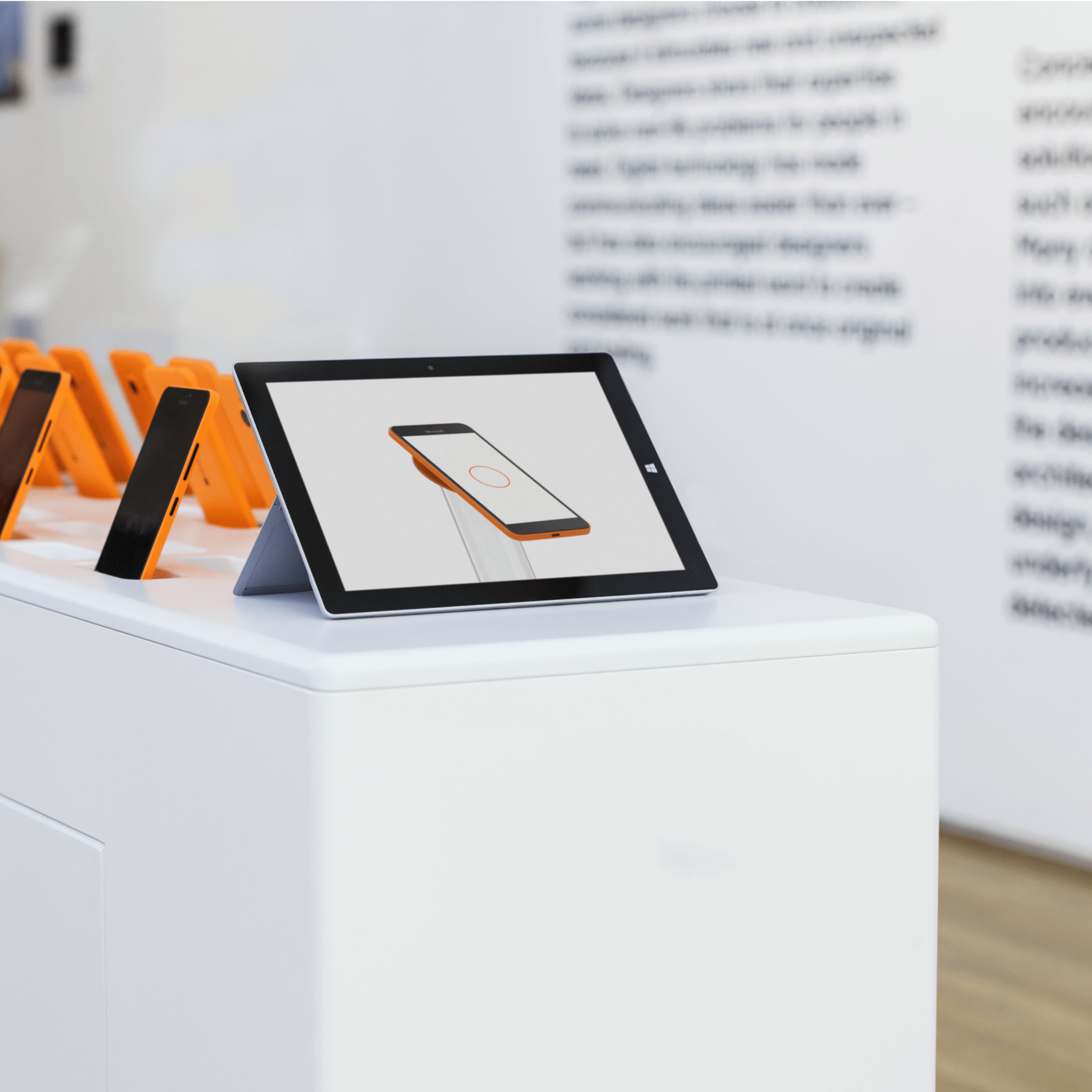 A Microsoft Surface tablet on a white pedestal displaying a Windows Phone image, with multiple Nokia Lumia smartphones in orange cases displayed upright in the background of a bright showroom.