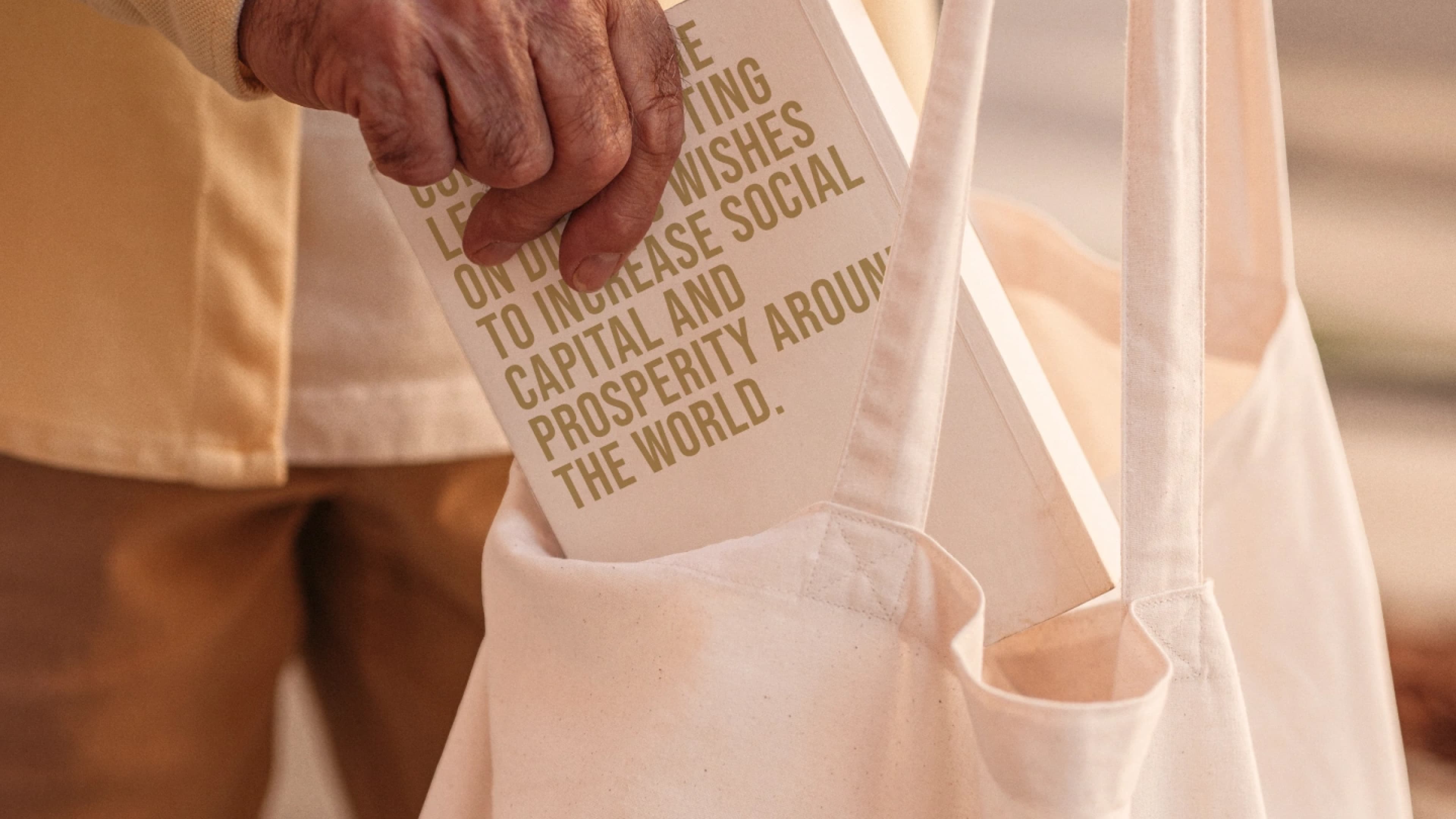 A person placing a printed card or booklet into a beige canvas tote bag. The visible text reads: "…wishes to increase social capital and prosperity around the world." — likely Maradona Foundation branded material.