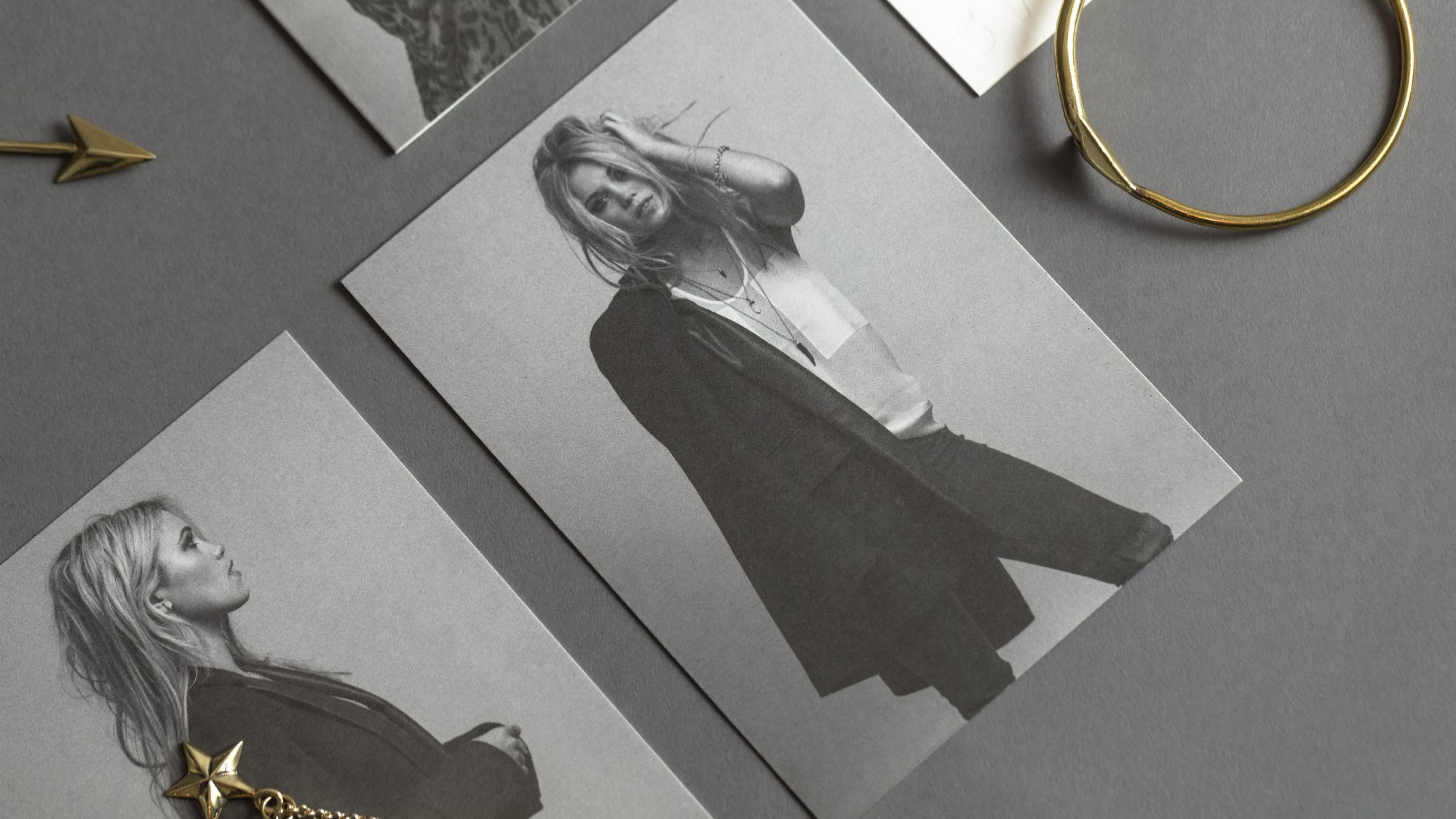 Monochrome illustrated portrait cards of a blonde woman with loose hair wearing a dark jacket, arranged on a grey surface alongside a gold bangle and star charm.