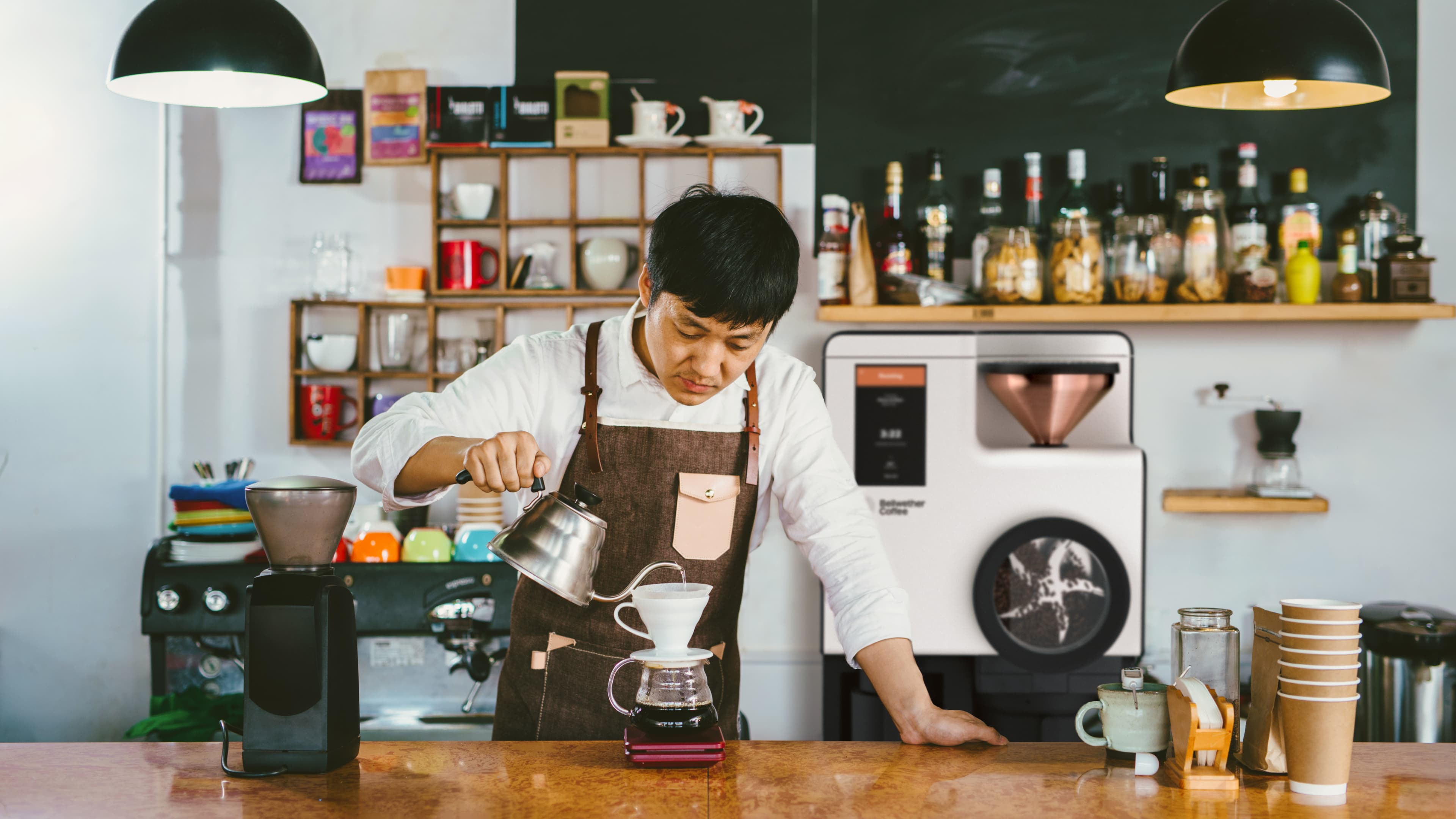 Barista in a white shirt and brown apron carefully pouring hot water from a gooseneck kettle into a pour-over dripper on a digital scale, behind a café counter with equipment and shelves of cups and bottles in the background.