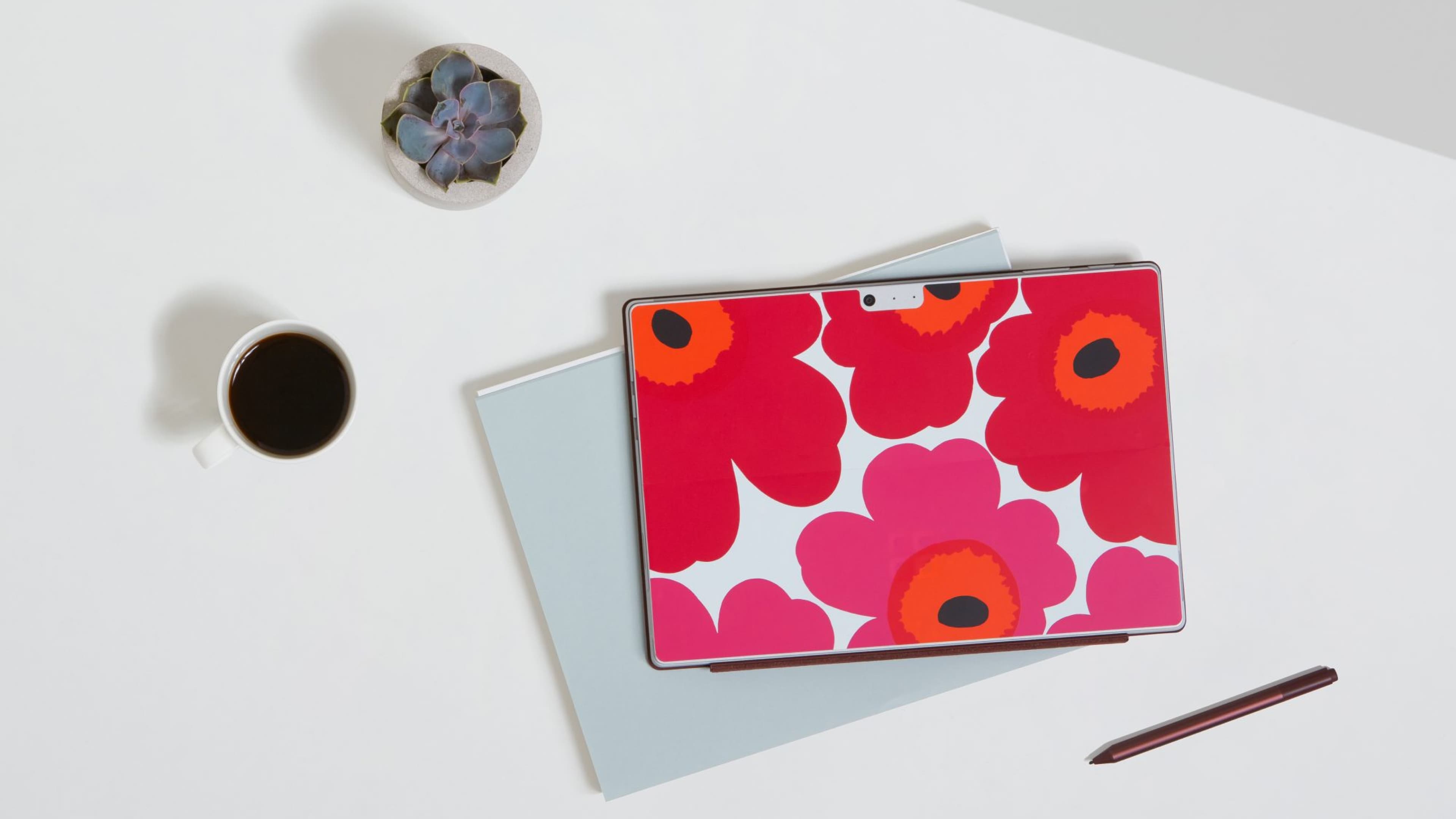 Microsoft Surface laptop with Marimekko Unikko red and pink floral pattern cover, displayed closed on a light blue folder on a white desk, alongside a succulent, a cup of black coffee, and a stylus pen.