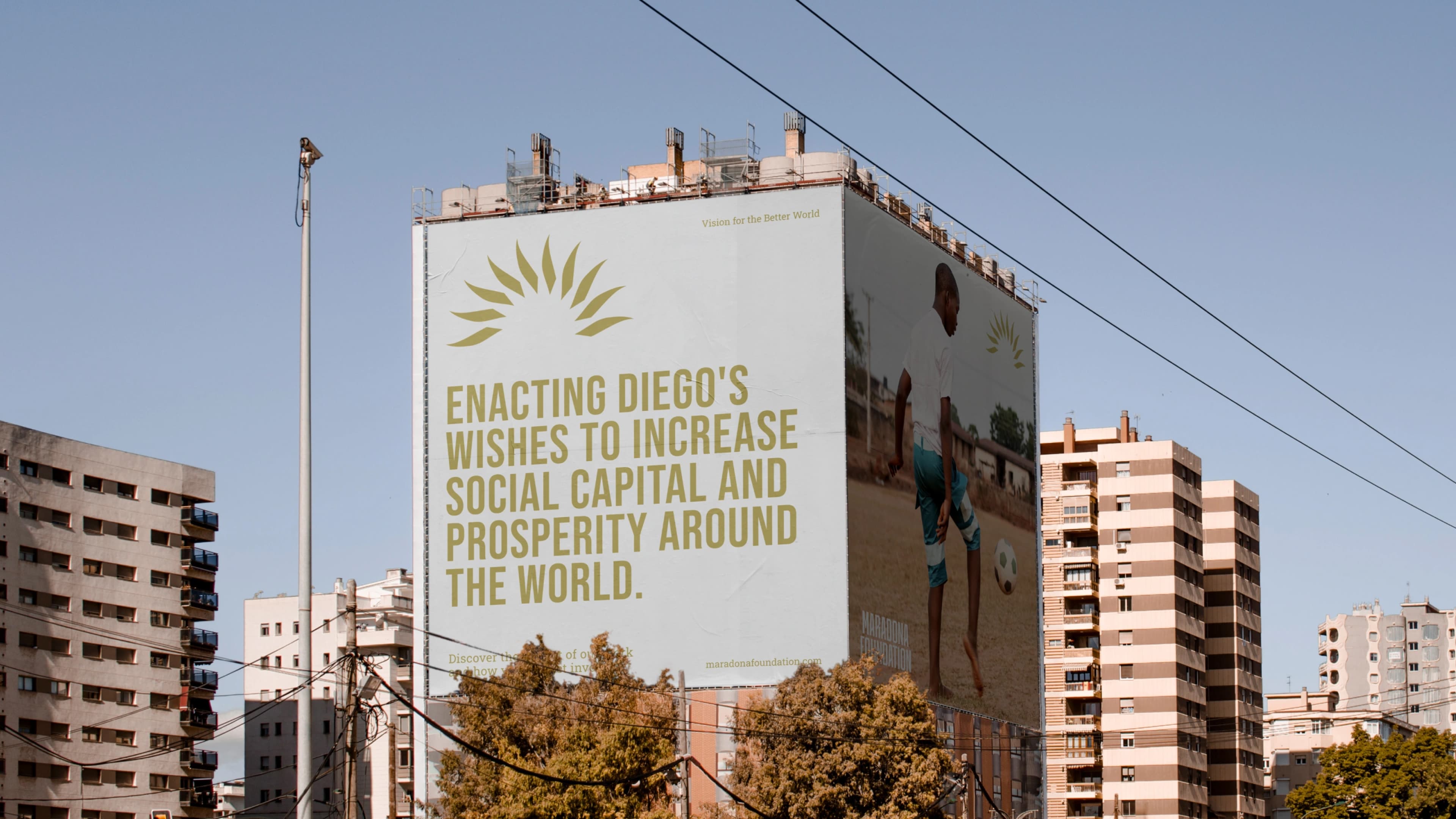 Large billboard on a building facade displaying a Maradona Foundation message reading "Enacting Diego's wishes to increase social capital and prosperity around the world," alongside an image of a child playing soccer in a sandy area. Surrounding urban area features tall residential buildings.