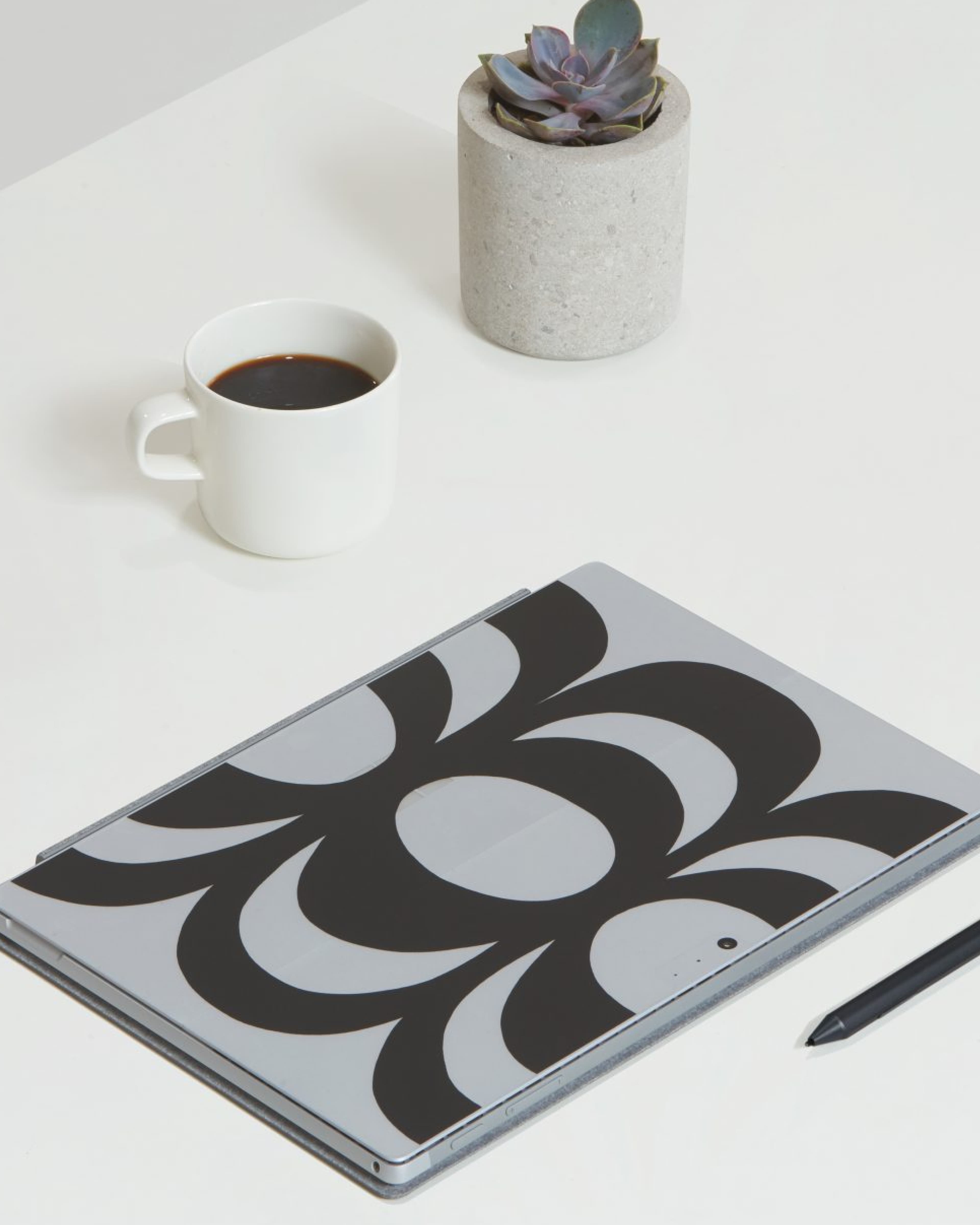A closed Microsoft Surface laptop with a bold Marimekko black-and-white swirl pattern skin, on a white desk beside a black coffee in a white mug, a concrete pot with a succulent, and a Surface Pen.