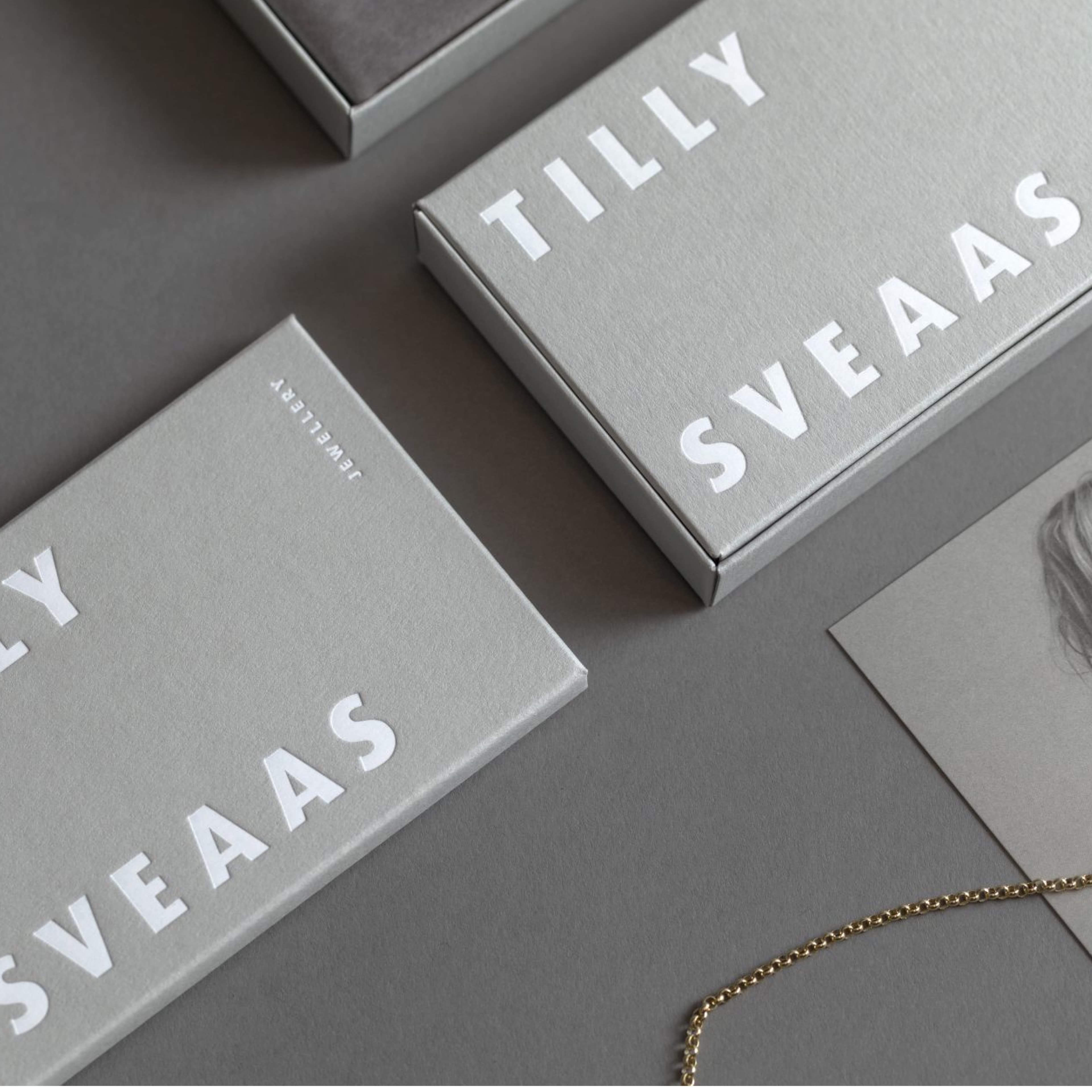 Gray Tilly Sveaas Jewellery gift boxes with bold white branding, displayed alongside a gold chain necklace and a partial card on a gray surface.