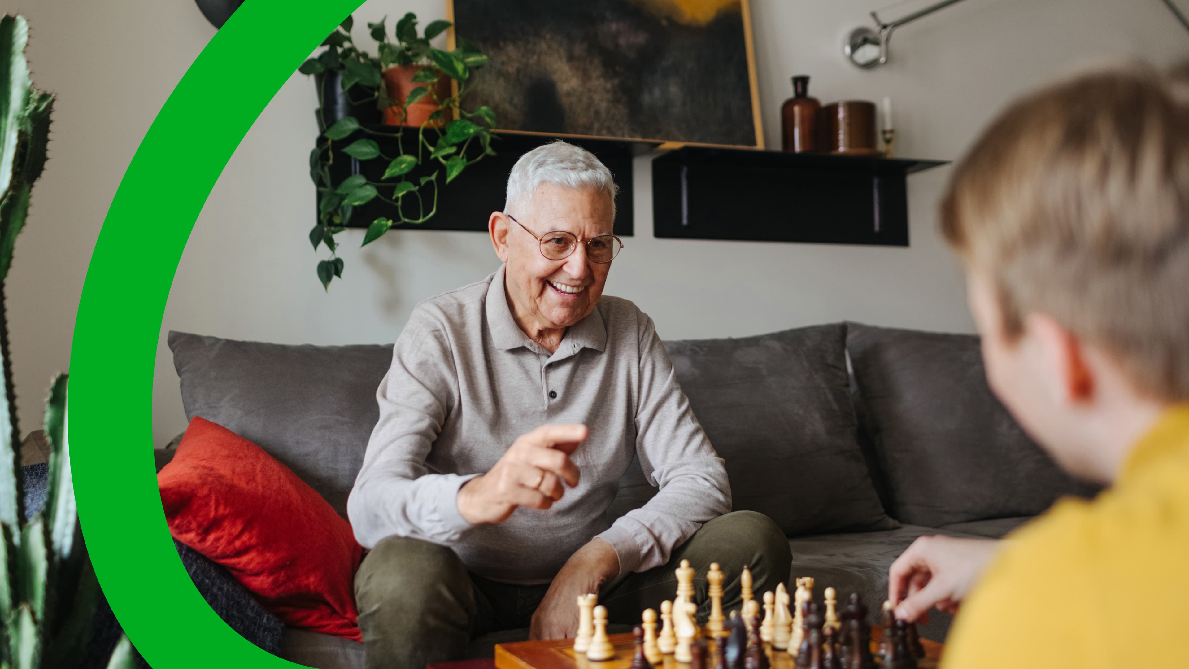 Elderly man playing chess with a younger person in a cozy living room with plants, a gray couch, red pillow, and artwork on the wall.