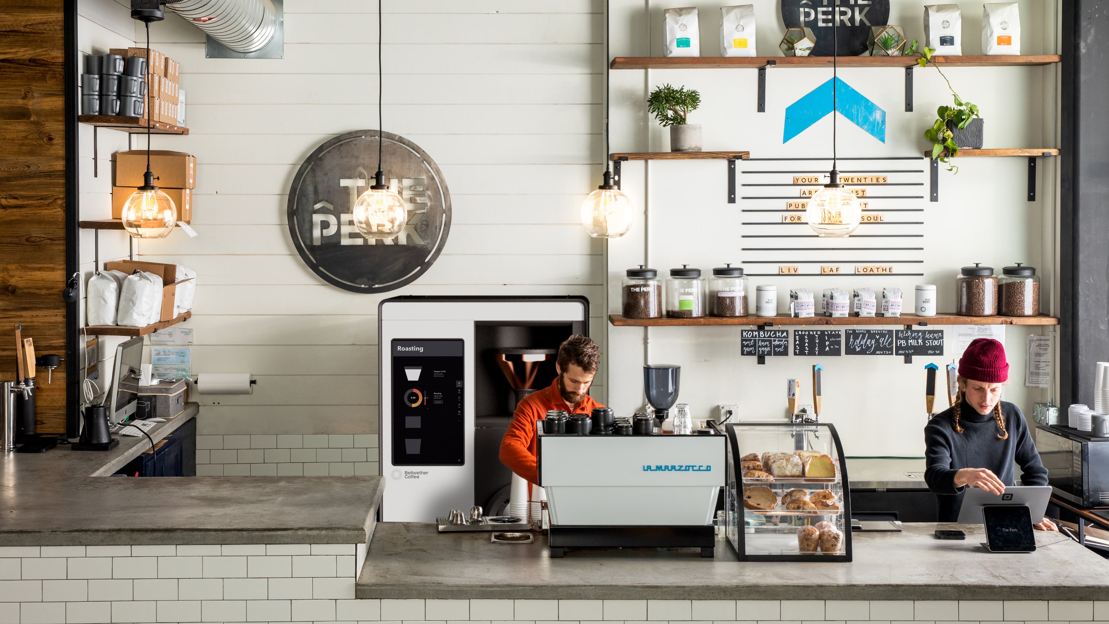 Interior of The Perk coffee shop featuring a Bellwether Coffee roaster and La Marzocco espresso machine on the counter, with a barista in an orange shirt and a staff member in a red beanie working at the register.