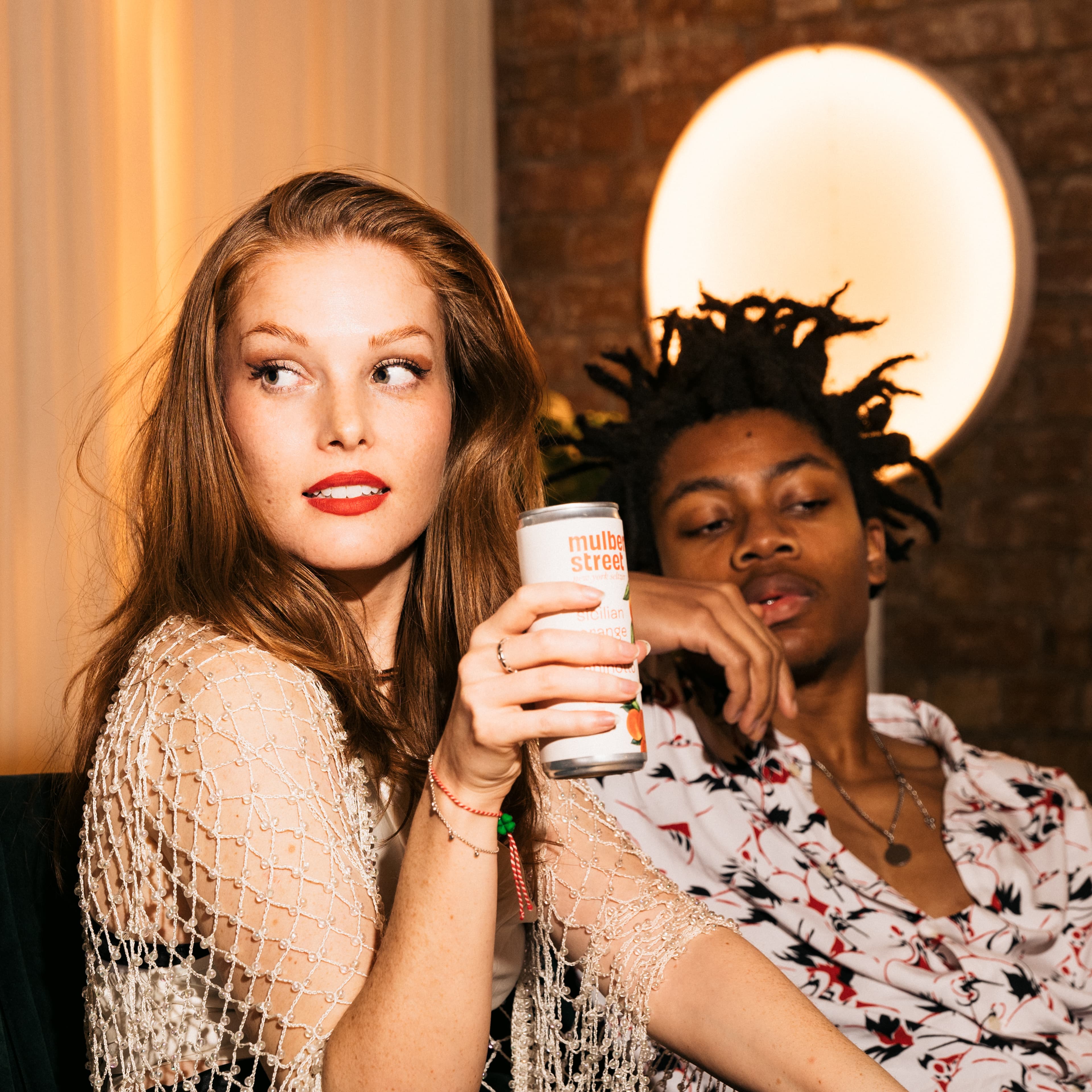 Two people sitting together at a warmly lit venue; one holds a Mulberry Street Sicilian orange canned drink and wears a beaded mesh top with red lipstick, the other wears a floral patterned shirt and has loc'd hair.