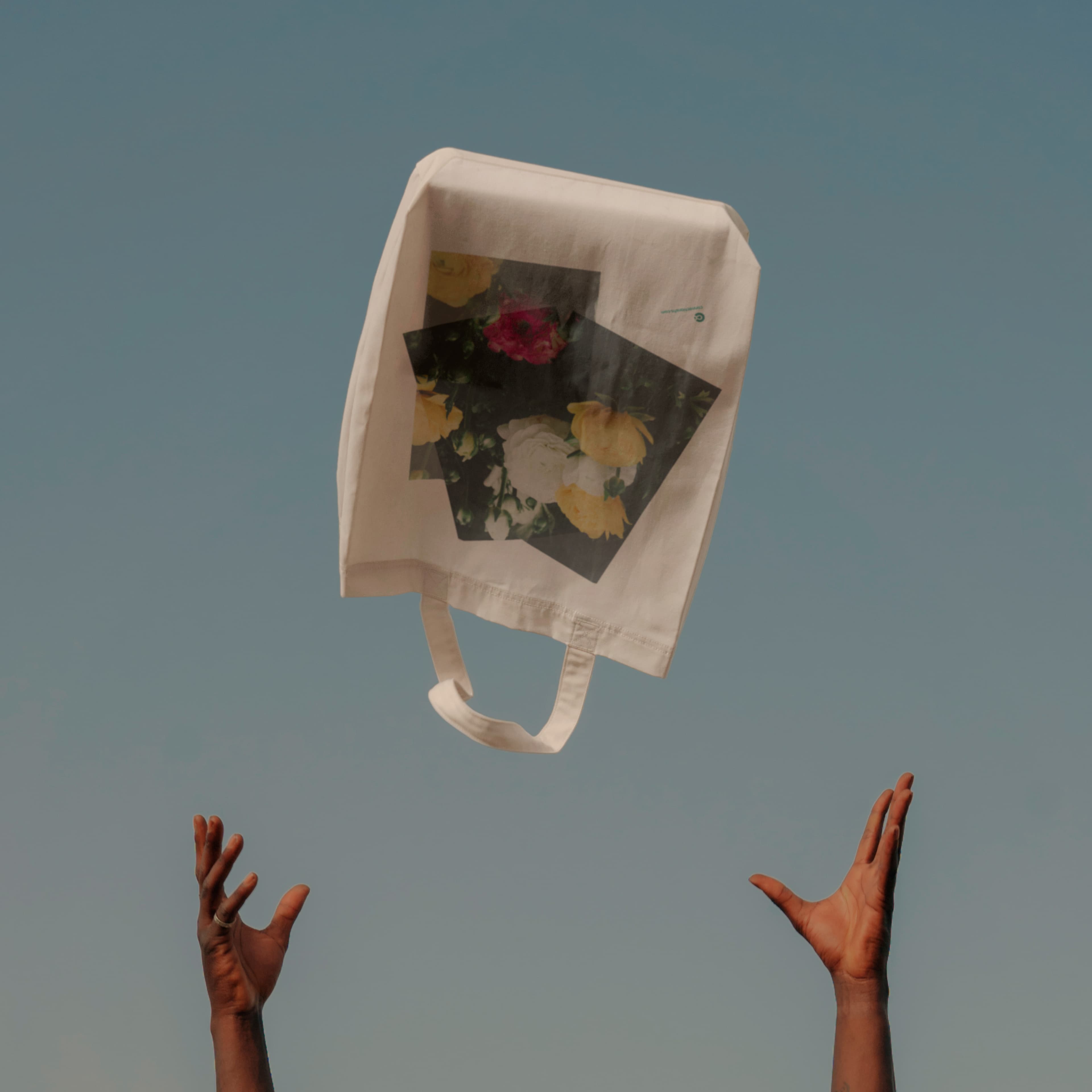 A white canvas tote bag with a photographic floral graphic print tossed in the air against a blue sky, with two hands reaching up toward it.