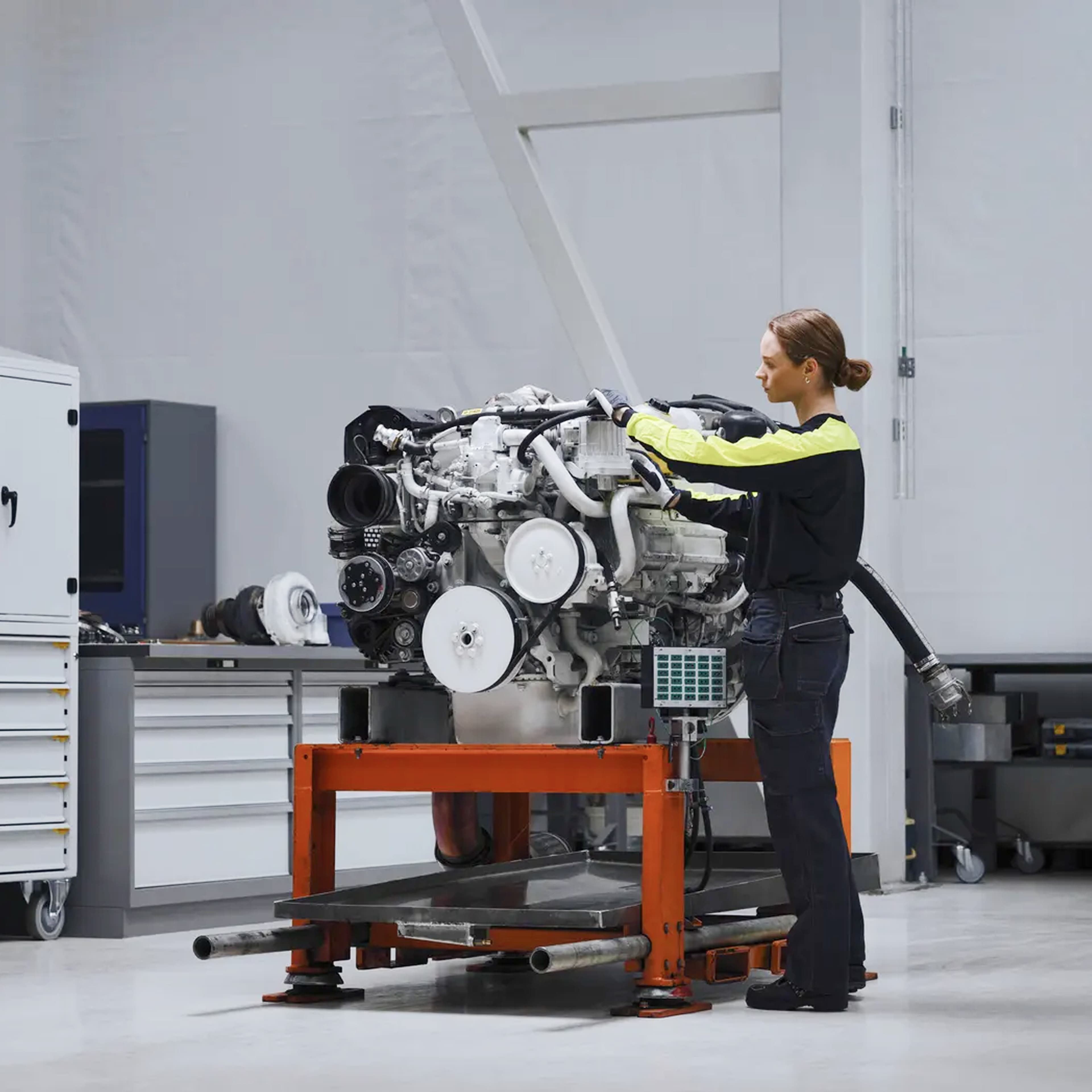 Picture of a woman working on an engine inside a factory wearing dark clothing.