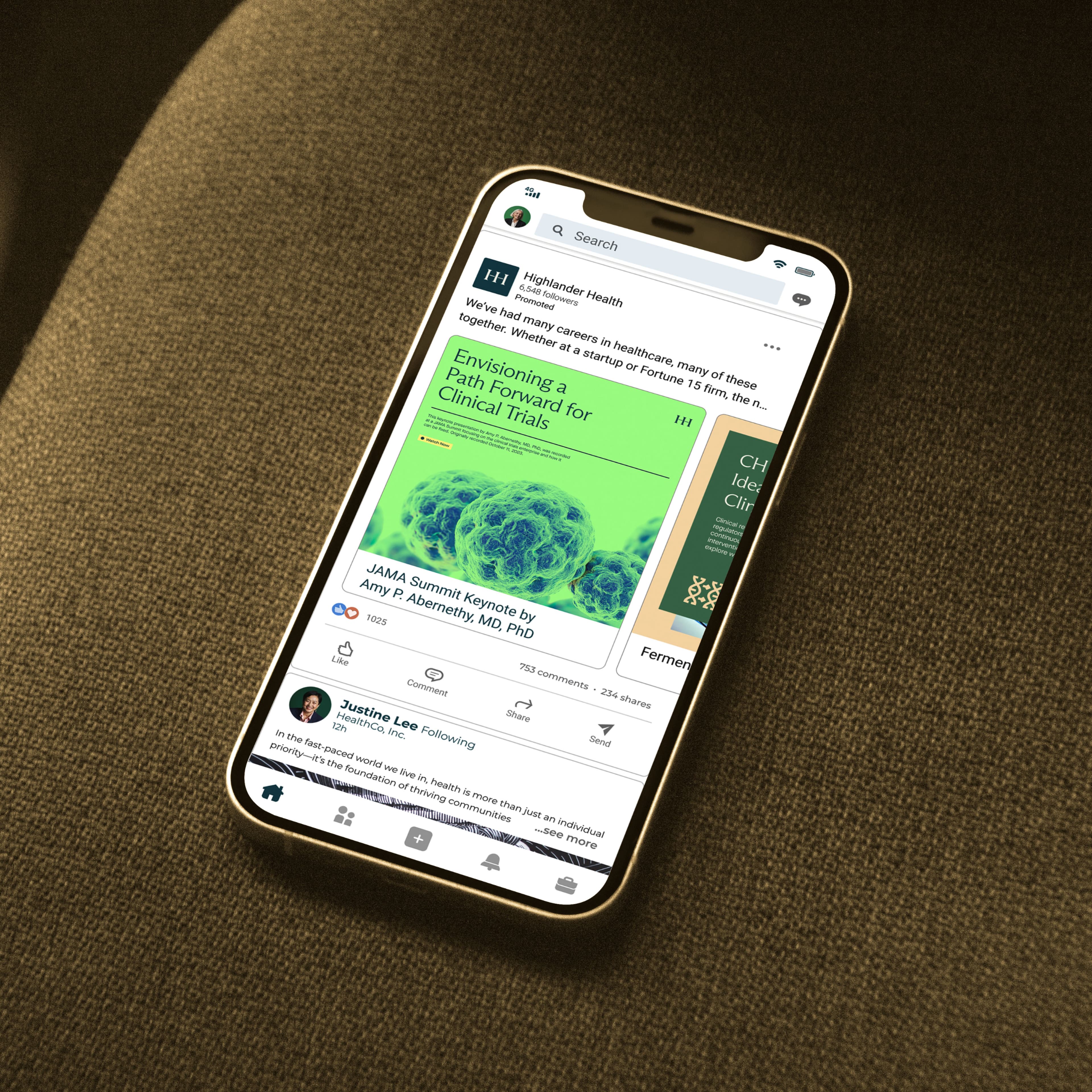 Smartphone on a brown fabric surface displaying a LinkedIn feed with a promoted post from Highlander Health featuring an article titled "Envisioning a Path Forward for Clinical Trials," a JAMA Summit keynote by Amy P. Abernethy, MD, PhD, shown with a green microscopy image.