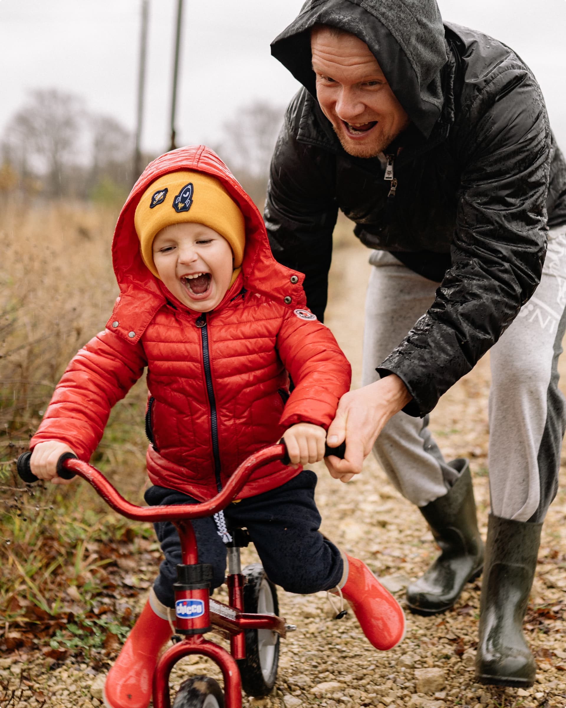 A laughing toddler in a red jacket and yellow hat rides a small red bicycle on a wet gravel path in the rain, while a man in a soaked black rain jacket helps steady the bike from behind.