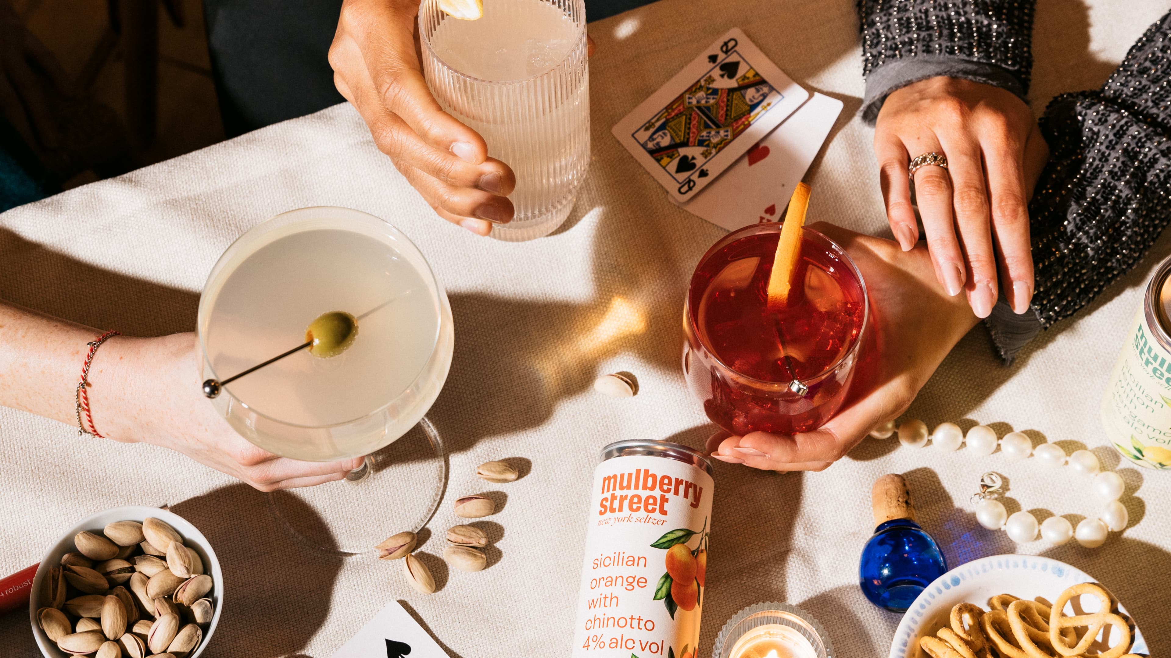 Two people holding cocktails — a martini with olive and a red drink with orange peel — over a table scattered with pistachios, mini pretzels, playing cards, a pearl necklace, and a Mulberry Street Sicilian Orange with Chinotto canned seltzer.