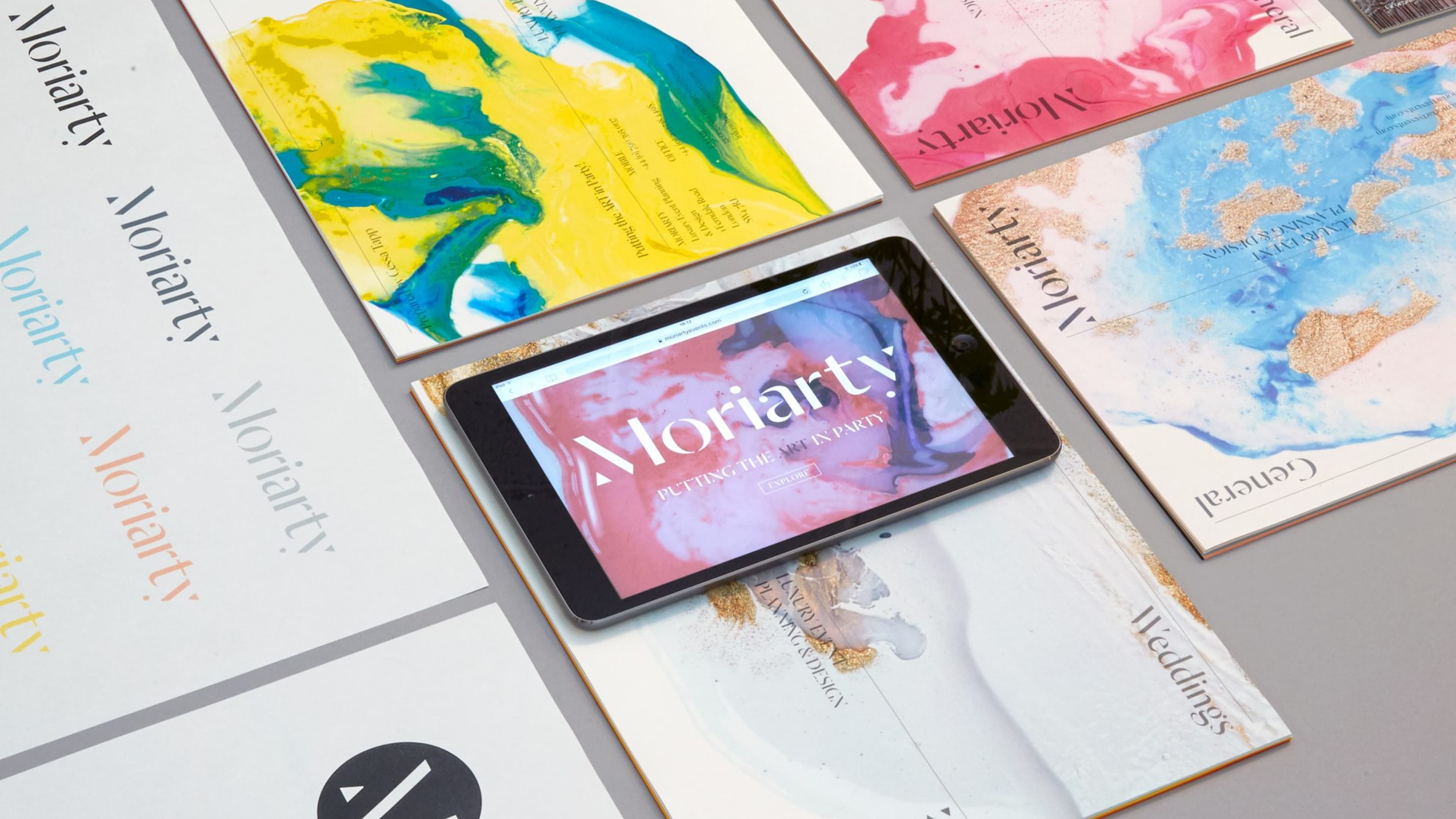 An arrangement of Moriarty print materials featuring colorful abstract covers and a tablet displaying a Moriarty webpage with similar art.
