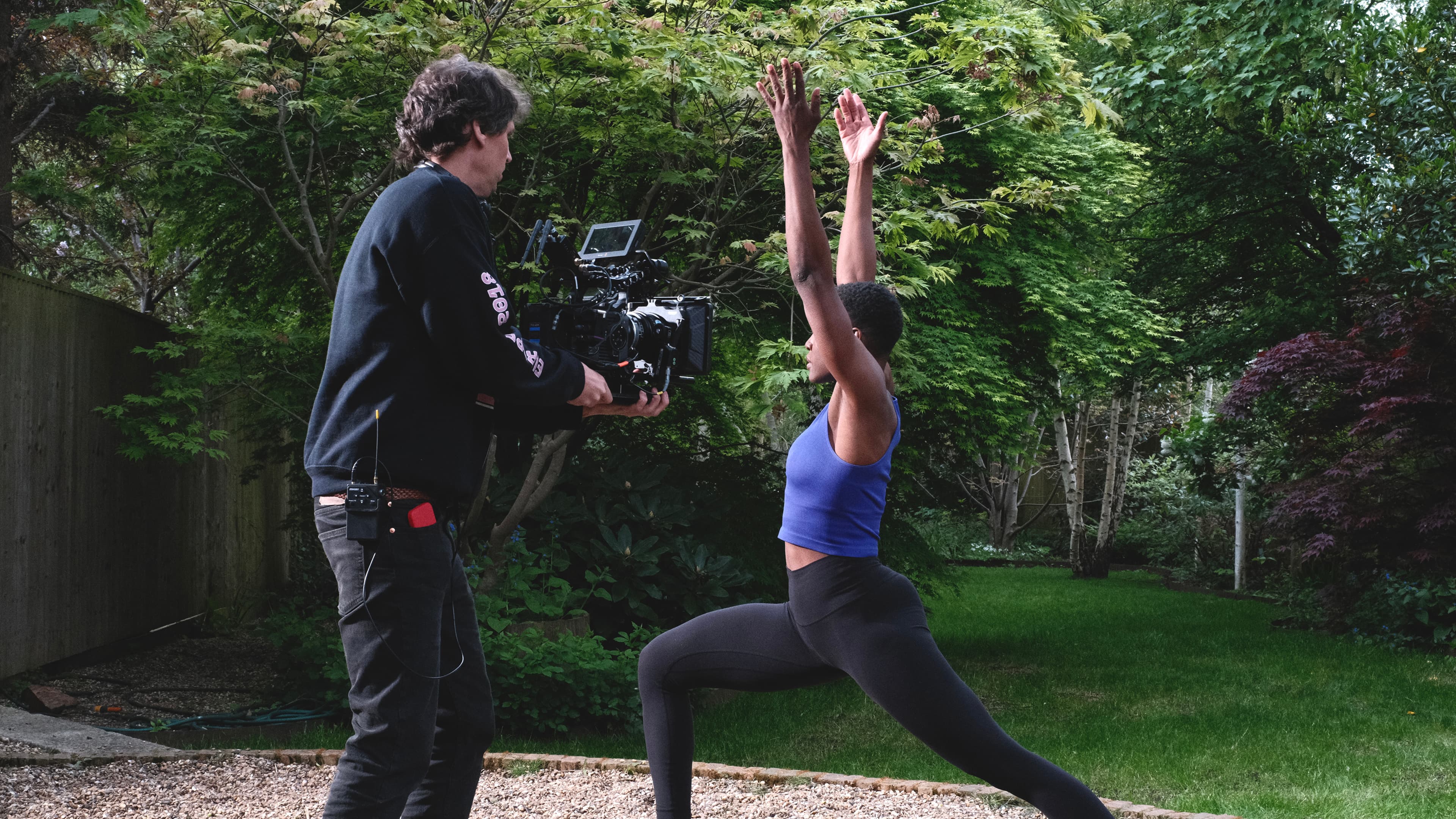 A filmmaker with a camera films a woman doing a yoga pose in a garden. She's wearing a blue top and black leggings, surrounded by green foliage.