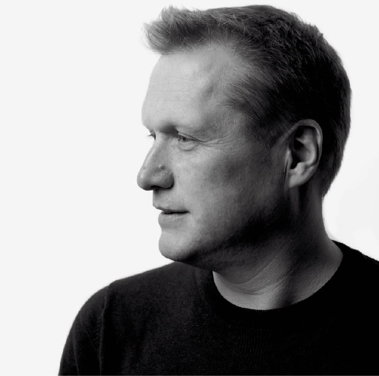 A man in profile view with a neutral expression, wearing a dark sweater, in a black and white portrait photo.