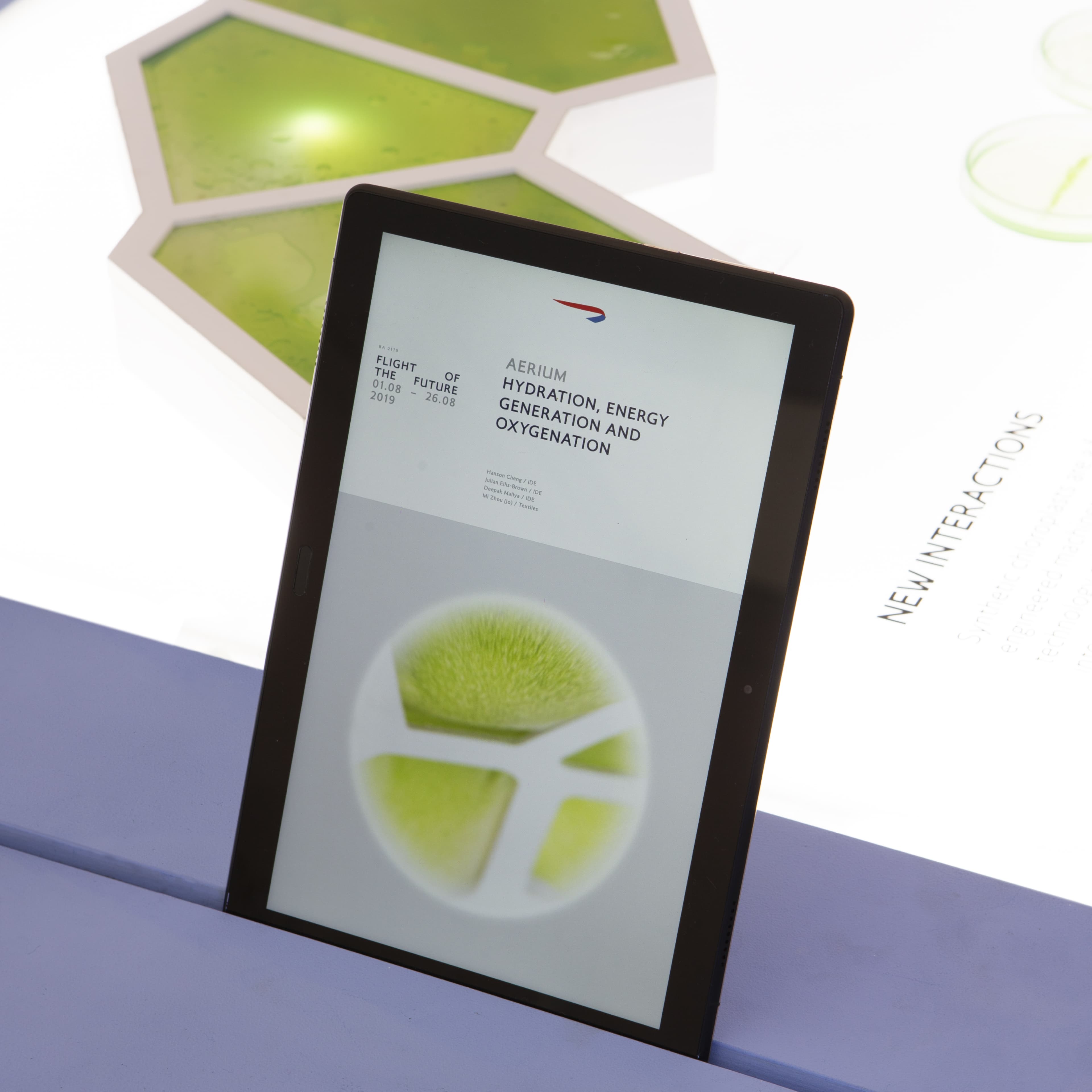 Tablet displaying the British Airways "Flight of the Future" exhibition project titled "Aerium: Hydration, Energy Generation and Oxygenation," dated 1–26 August 2019, with illuminated green hexagonal panels and "New Interactions" text visible in the background.