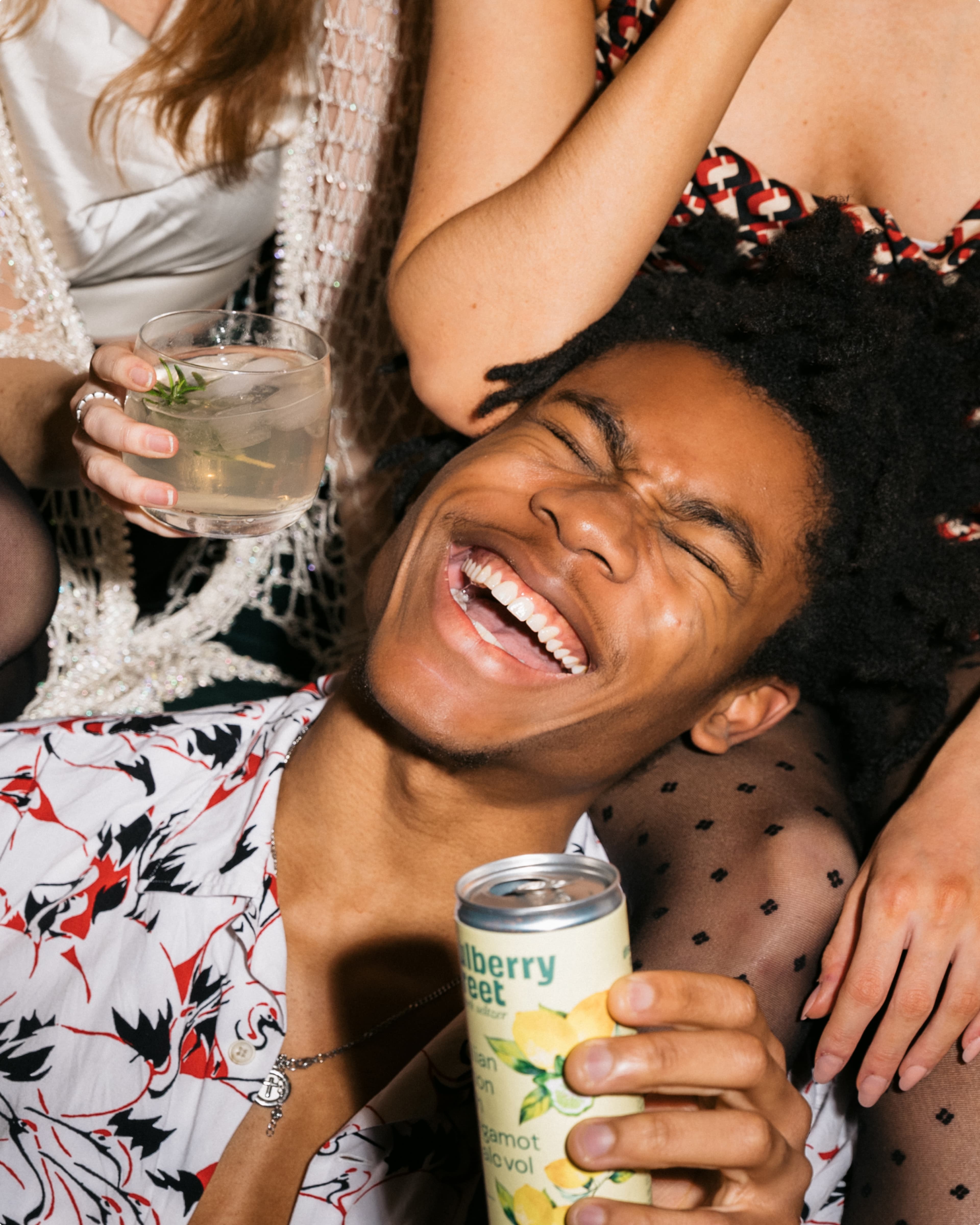A joyful person in a floral shirt holding a canned drink, surrounded by friends, one holding a cocktail, at a lively party.