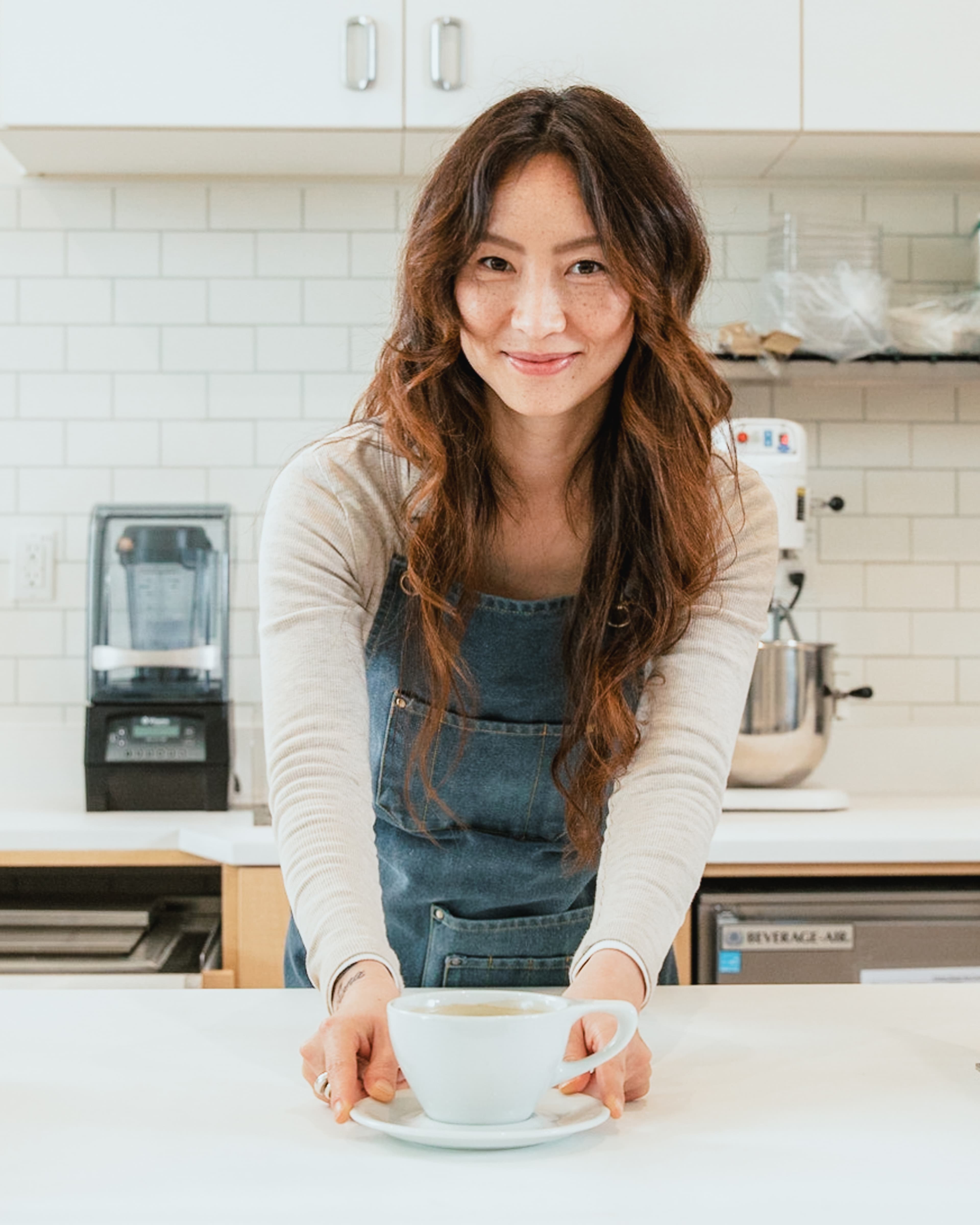 Smiling woman in a denim apron leaning over a white counter to present a cup of tea in a bright kitchen with subway tile walls and appliances in the background.