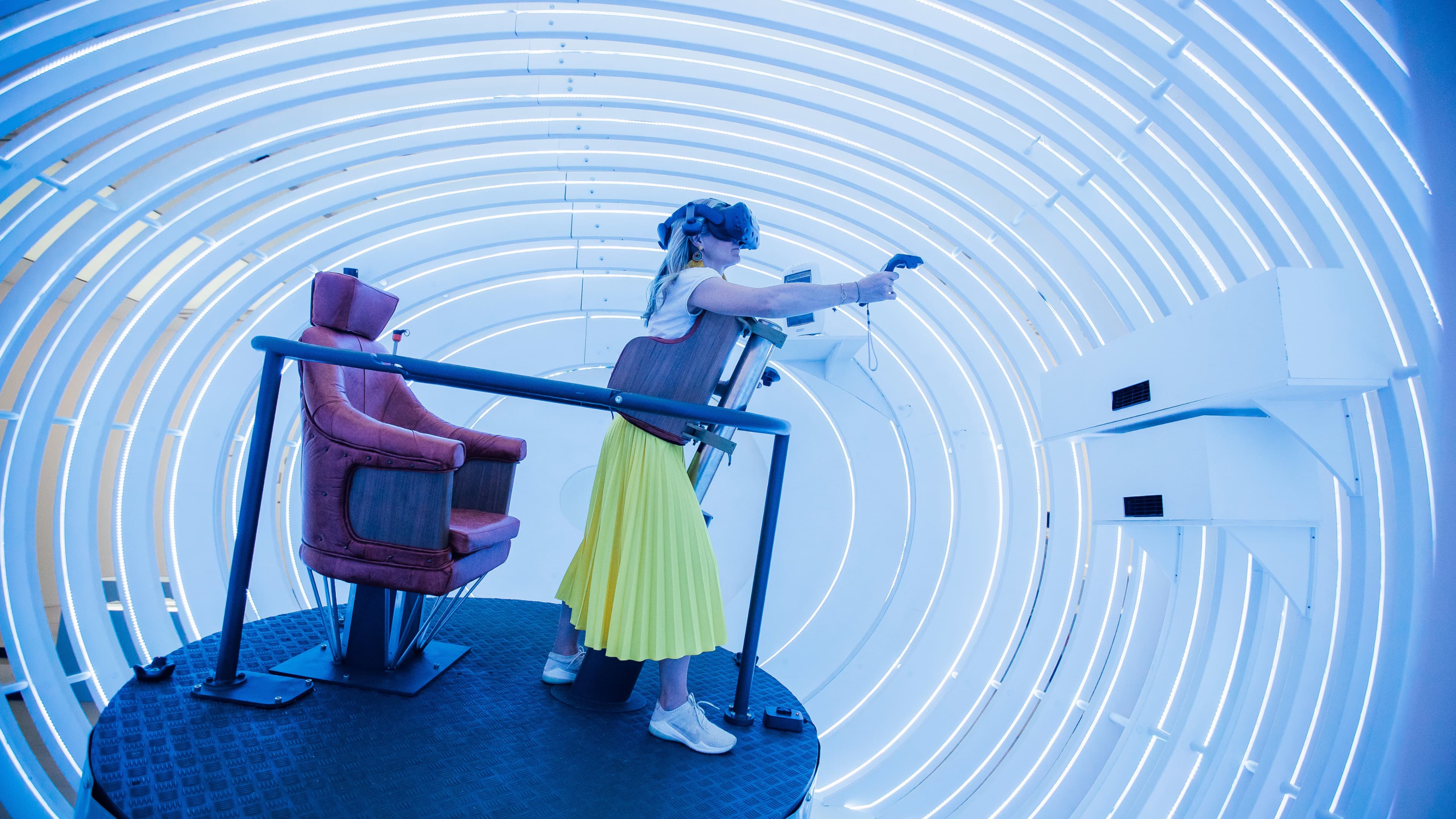 Person in VR headset in a futuristic, illuminated circular room with a yellow skirt and sneakers, interacting with a controller.