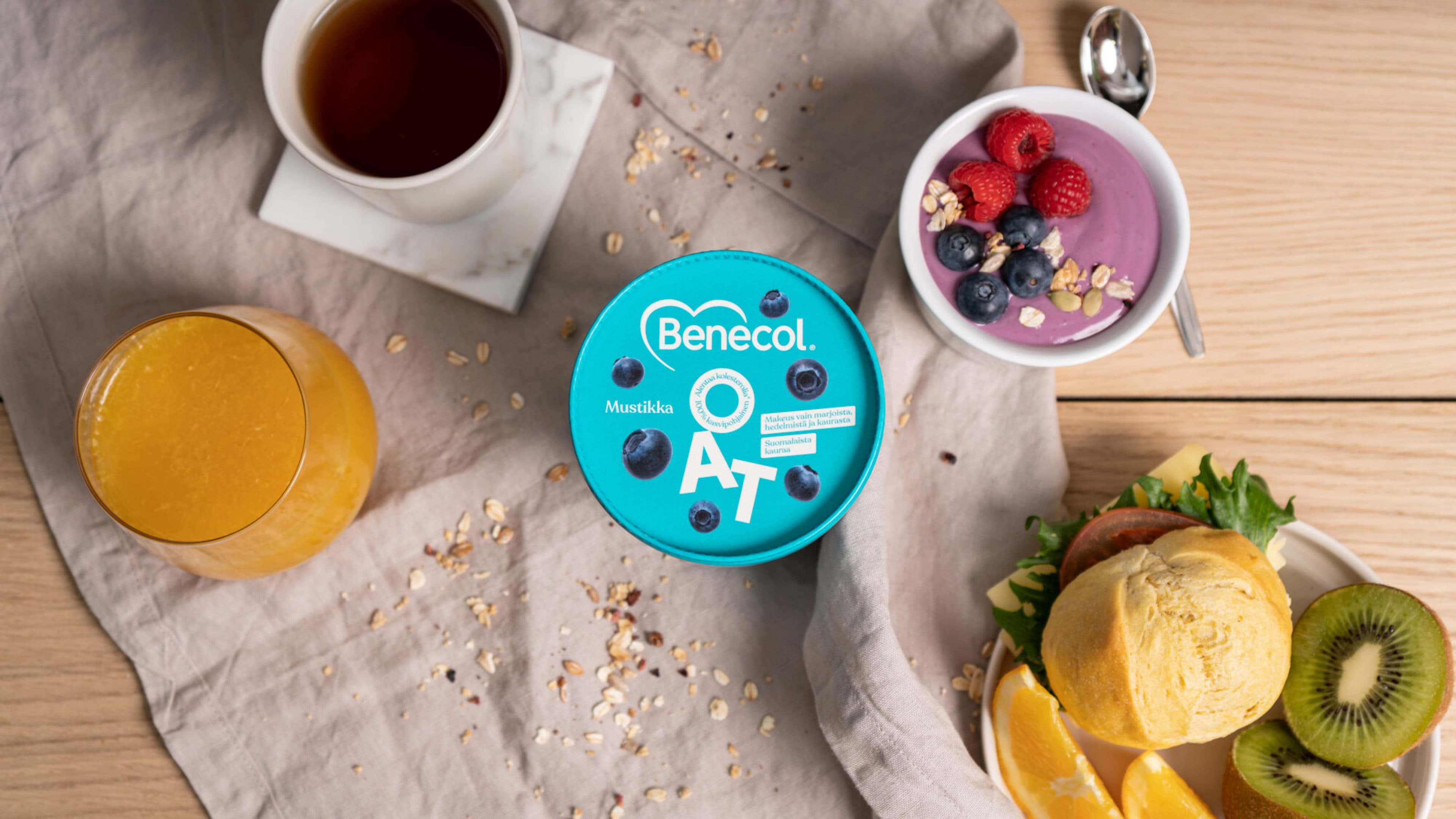 Breakfast spread with a cup of tea, orange juice, a Benecol Blueberry Oat container, a berry smoothie bowl topped with raspberries and blueberries, and a plate with a bread roll, kiwi, and orange slices.