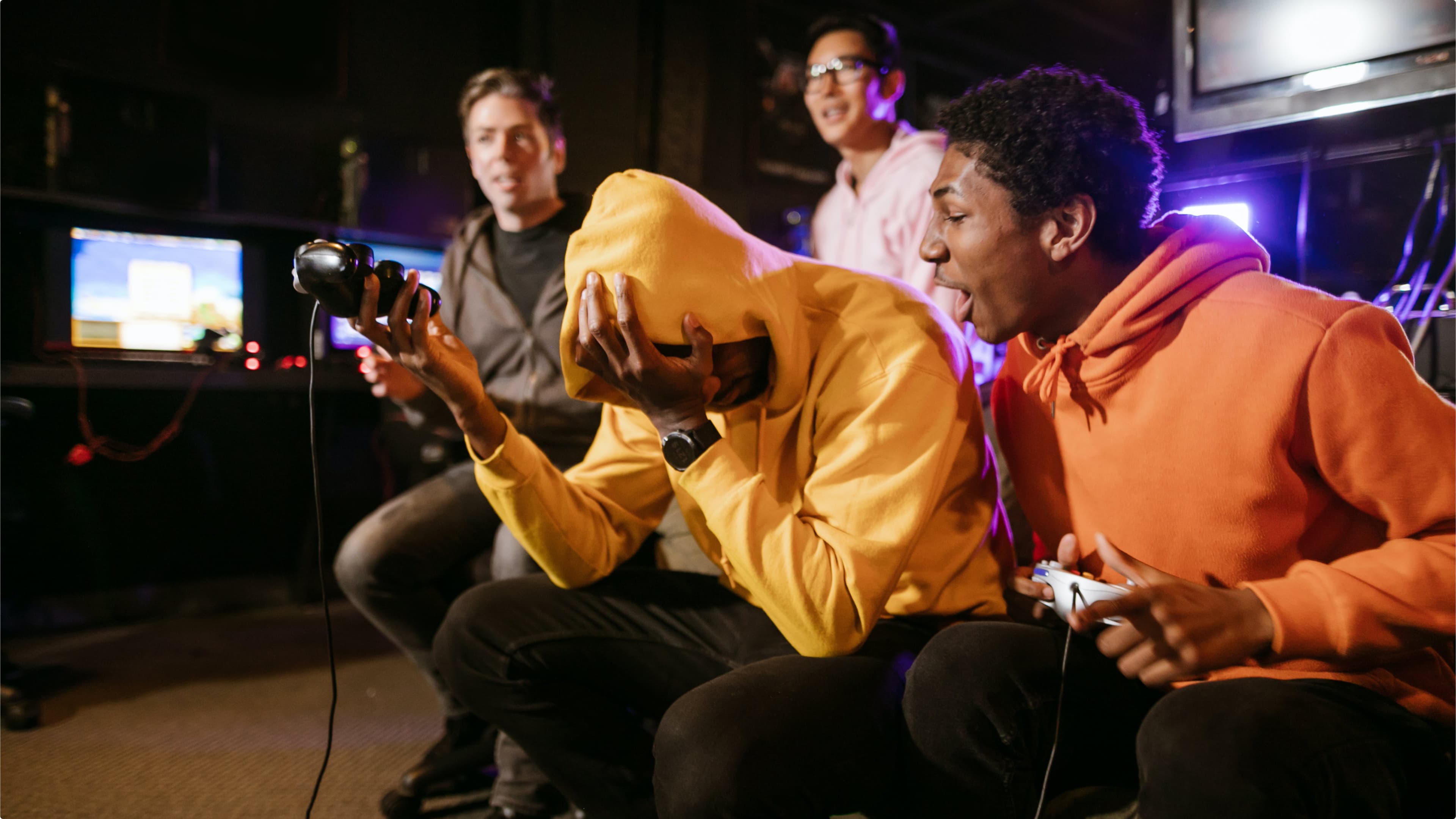Four friends at an indoor gaming session — one in a yellow hoodie buries their face in frustration, another in an orange hoodie reacts excitedly with a controller, while two others watch and smile in the background.