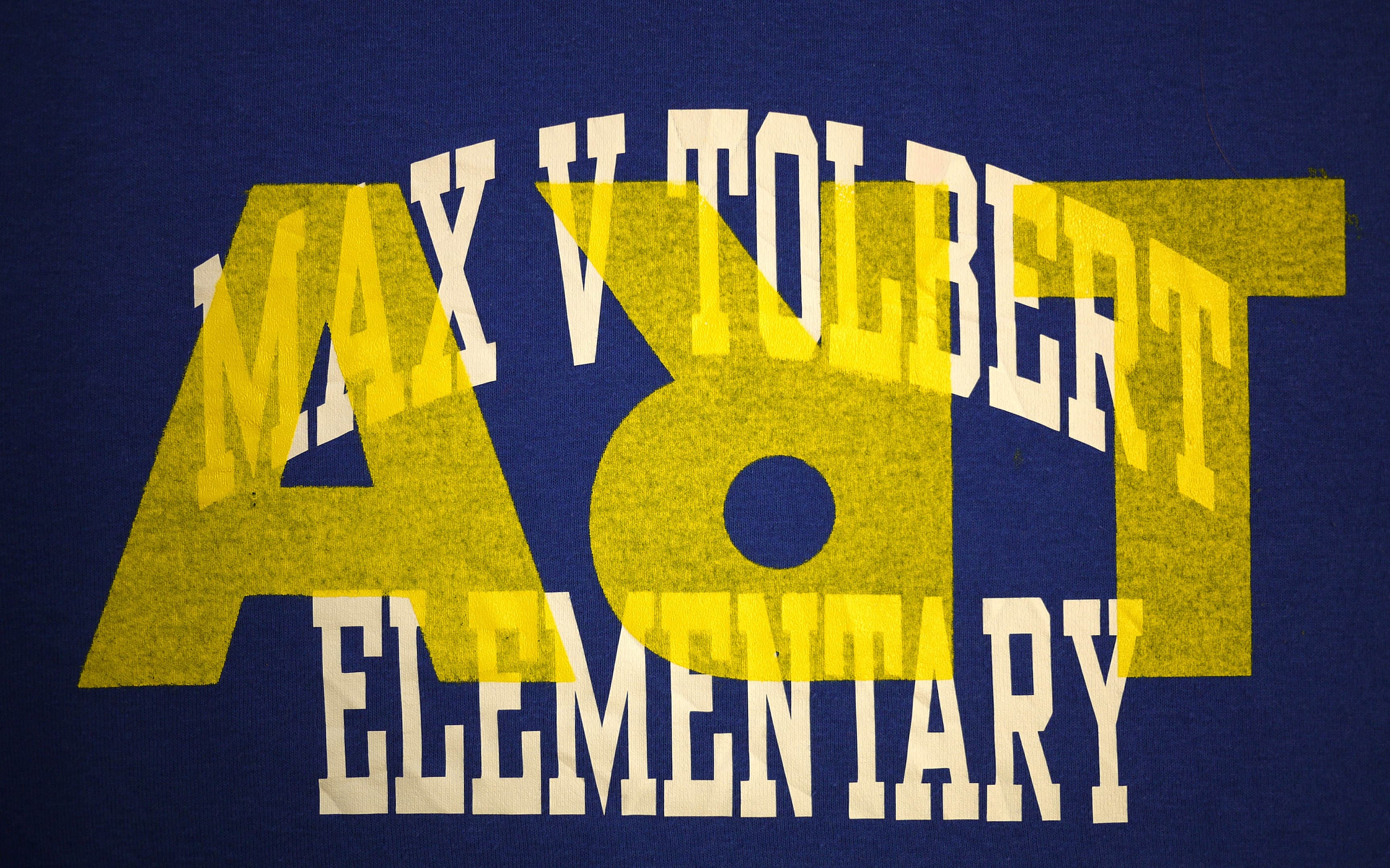 Blue T-shirt with large yellow "ART" letters overlaid with white arched text reading "Max V Tolbert Elementary."