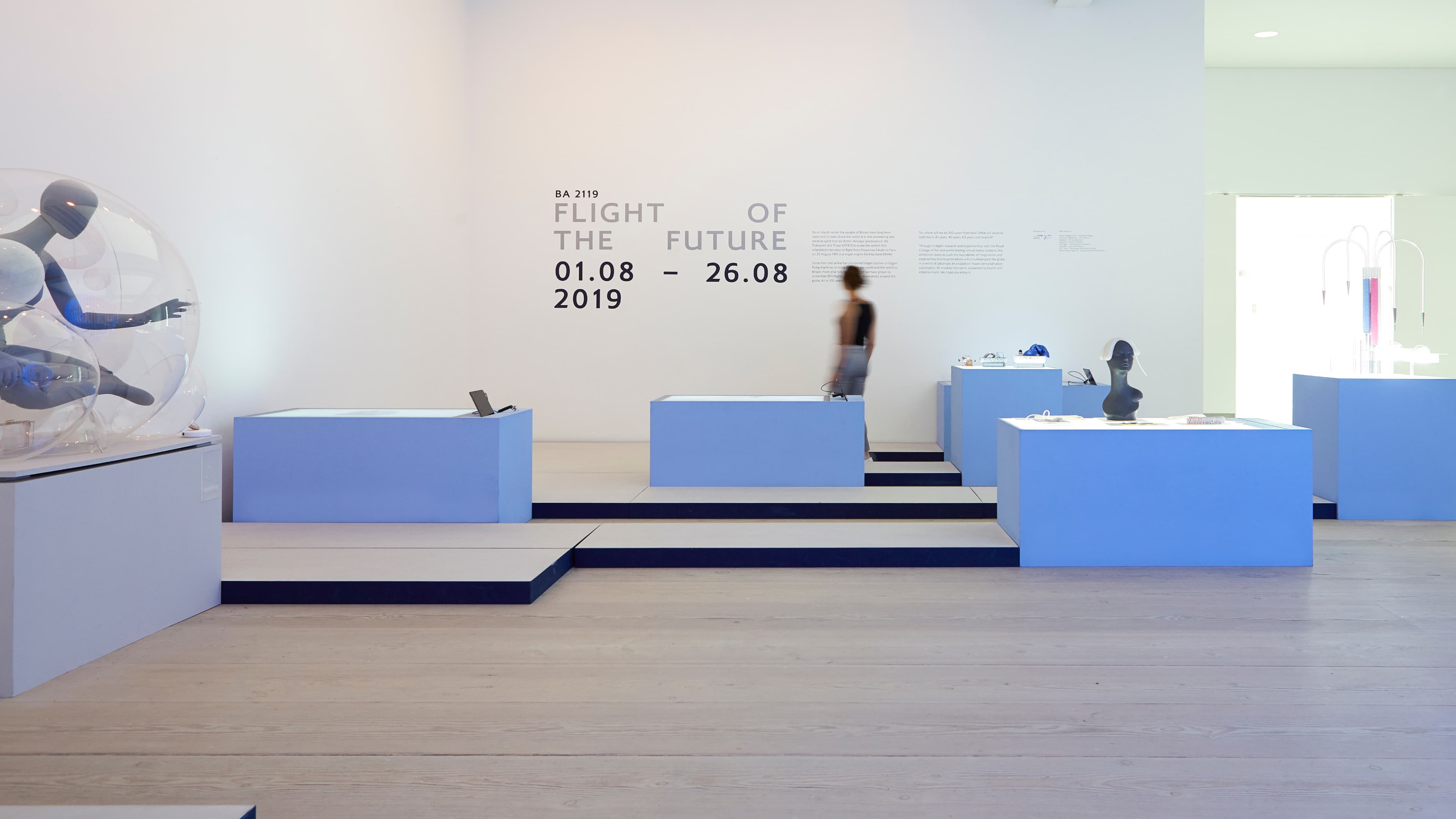 Interior of gallery exhibition "BA 2119: Flight of the Future" (1–26 August 2019), featuring blue display pedestals with objects, a transparent sphere encasing a mannequin figure, and a visitor walking through the space.