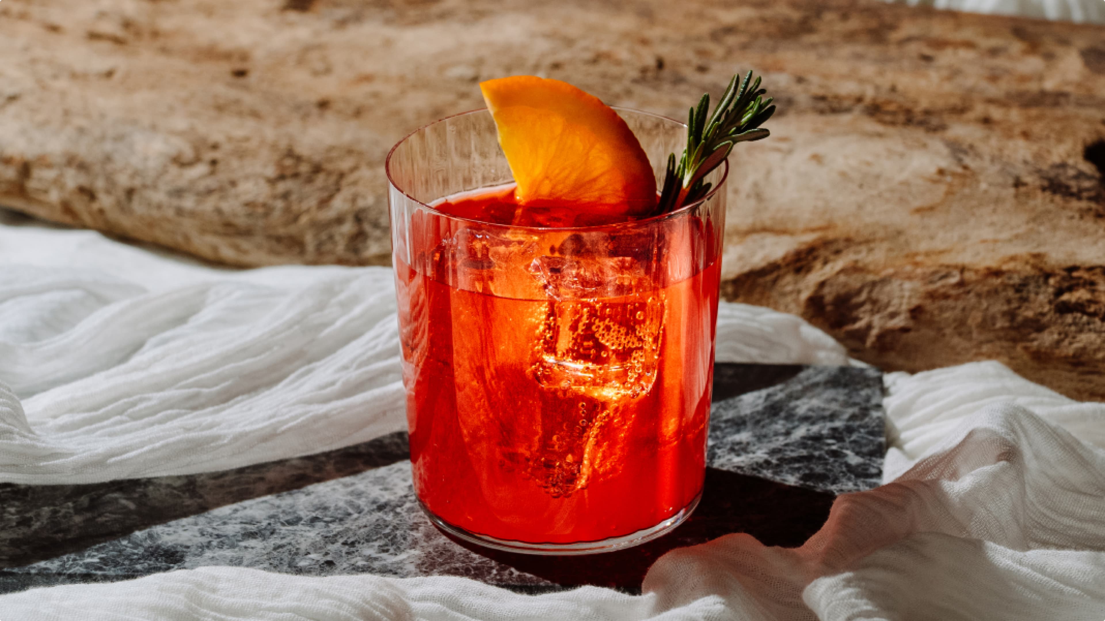 A vibrant Negroni cocktail in a rocks glass with ice, garnished with an orange slice and a sprig of rosemary, set on a marble surface with a driftwood background.