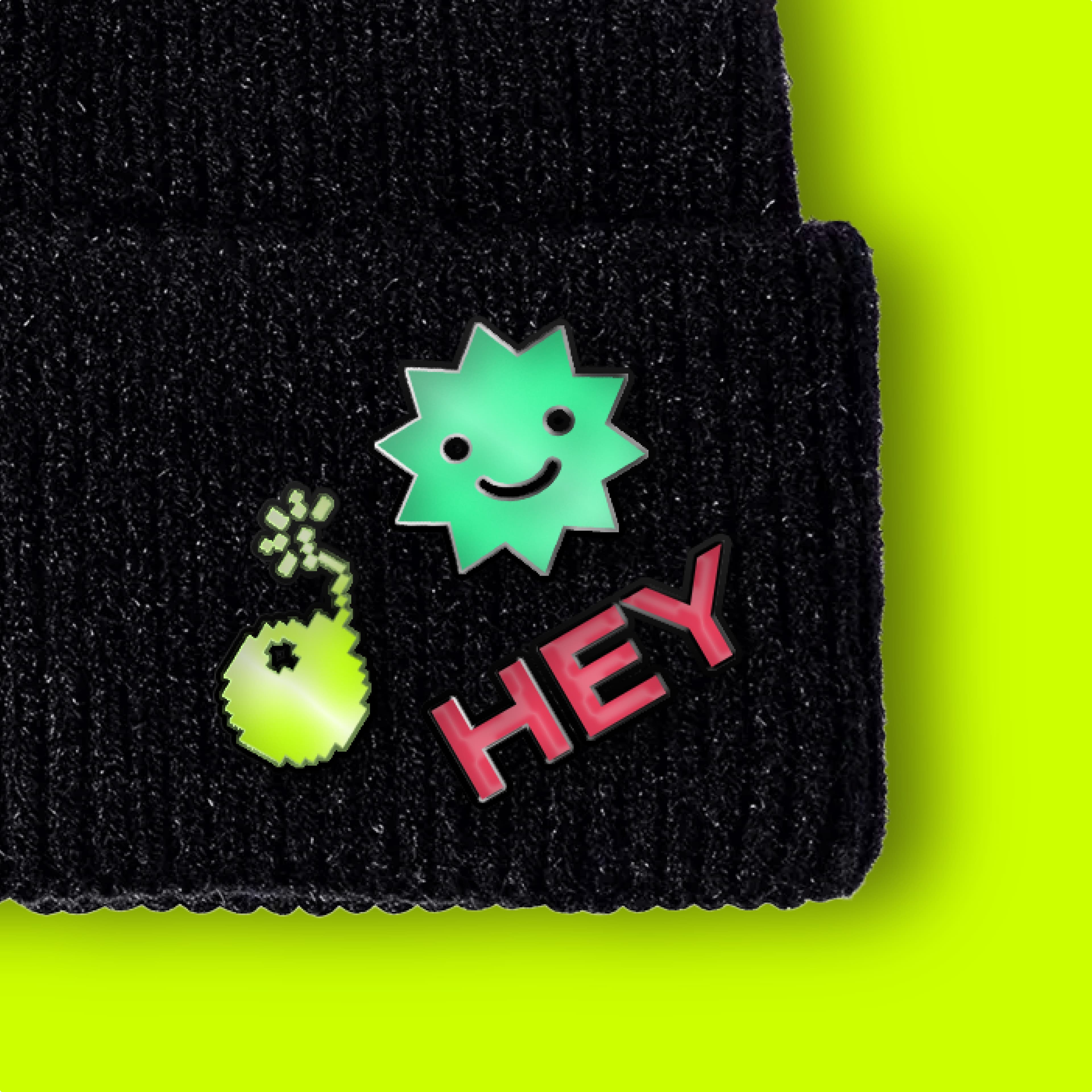 Black knitted beanie with green smiley star, small green blob, and red 'HEY' pin against a bright yellow-green background.