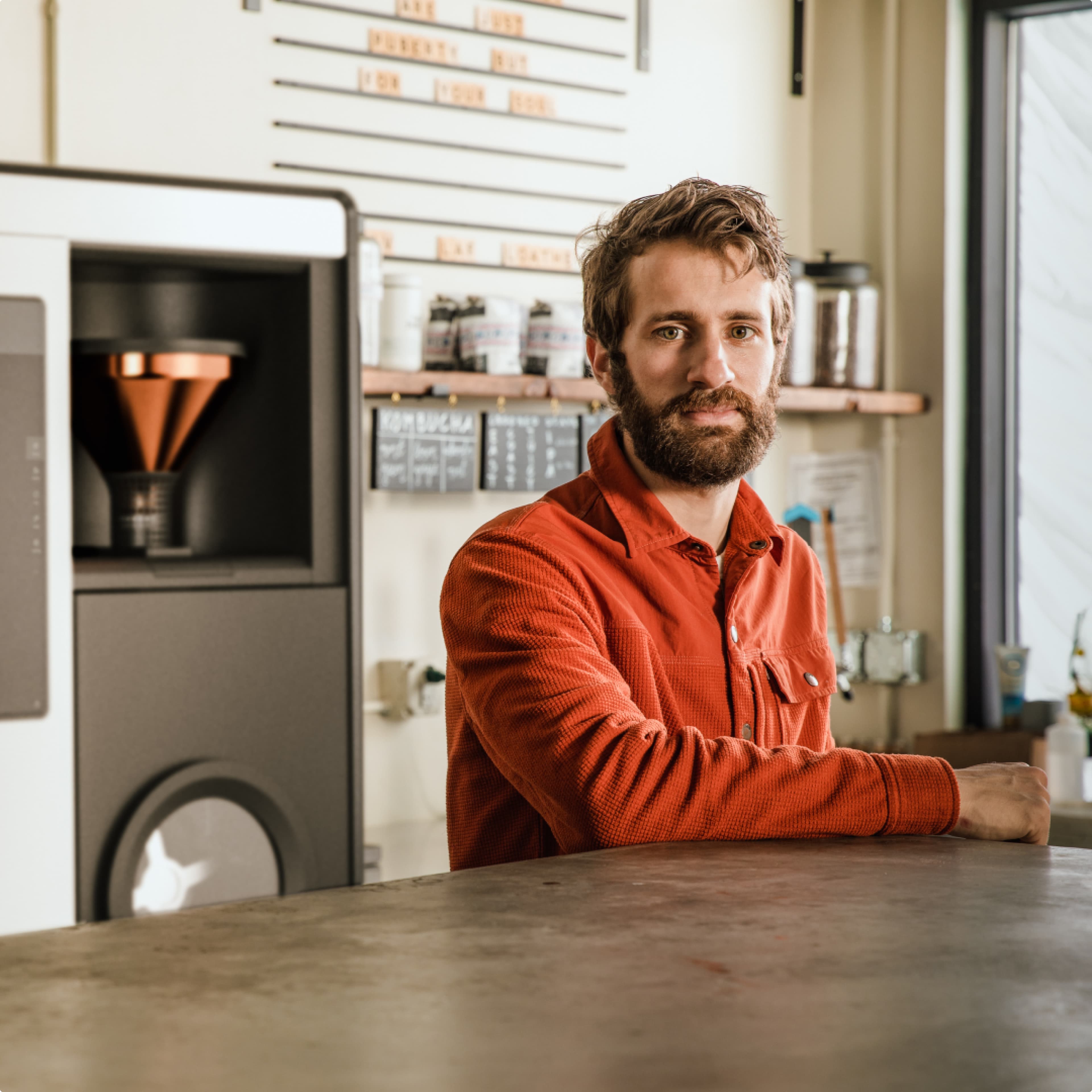 A man with a beard in an orange shirt leans on a counter in a cafe, with a coffee machine and shelves in the background.