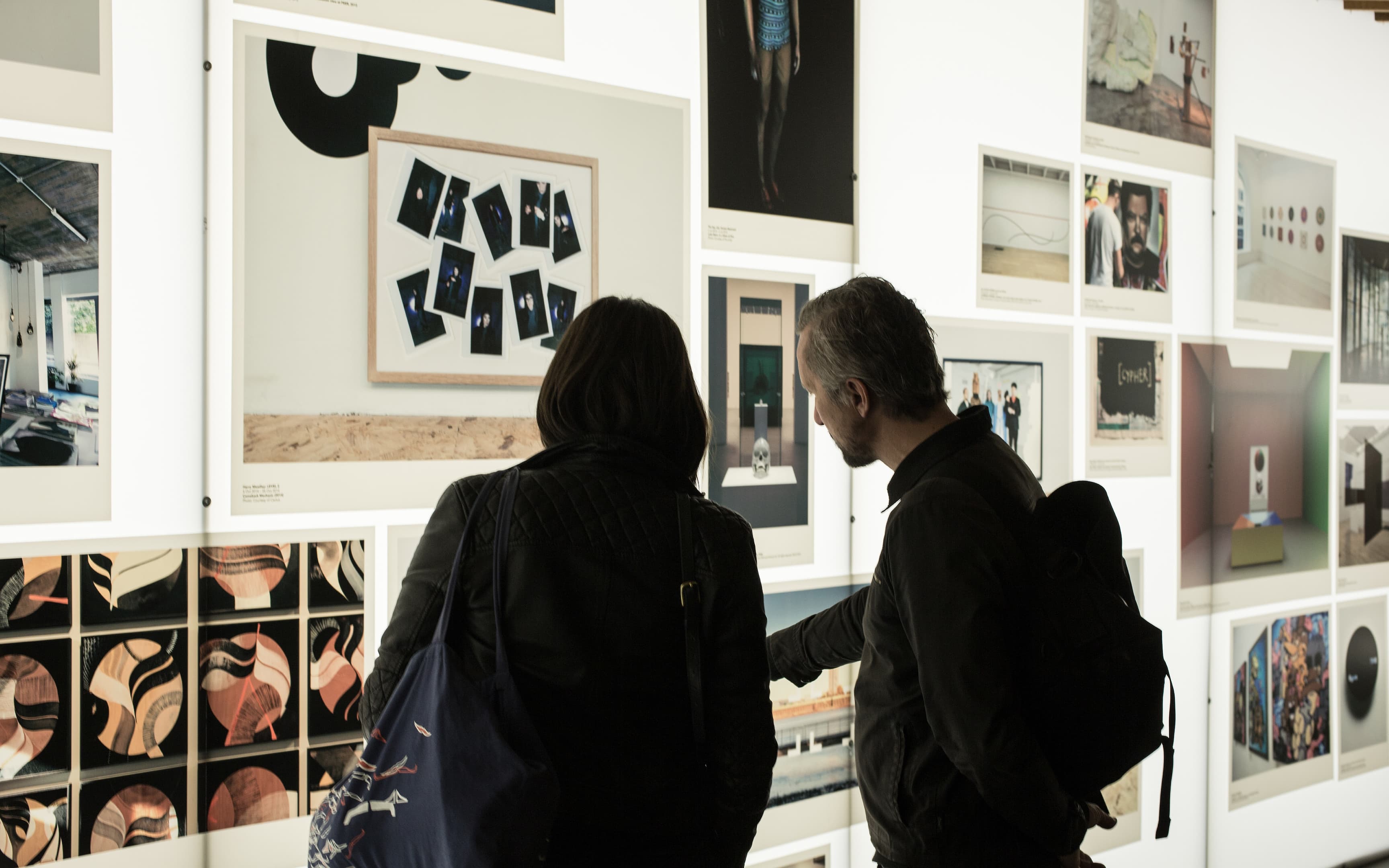 Two people in dark jackets observe a gallery wall with various framed artworks, including photos and abstract designs, under bright lighting.