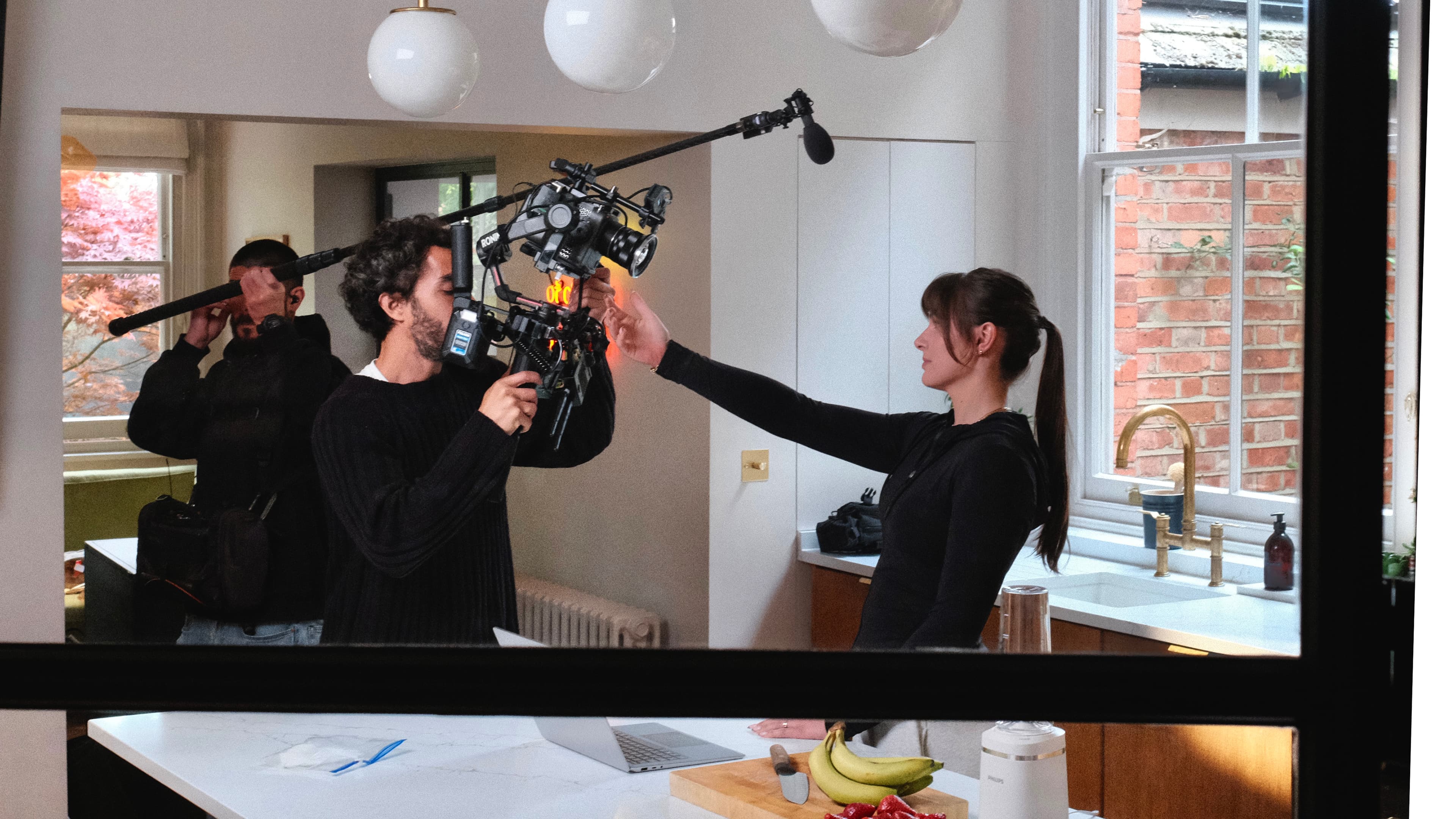 Film crew in a modern kitchen with a camera operator and a woman interacting. A man holds a boom mic near a window with brick wall view.