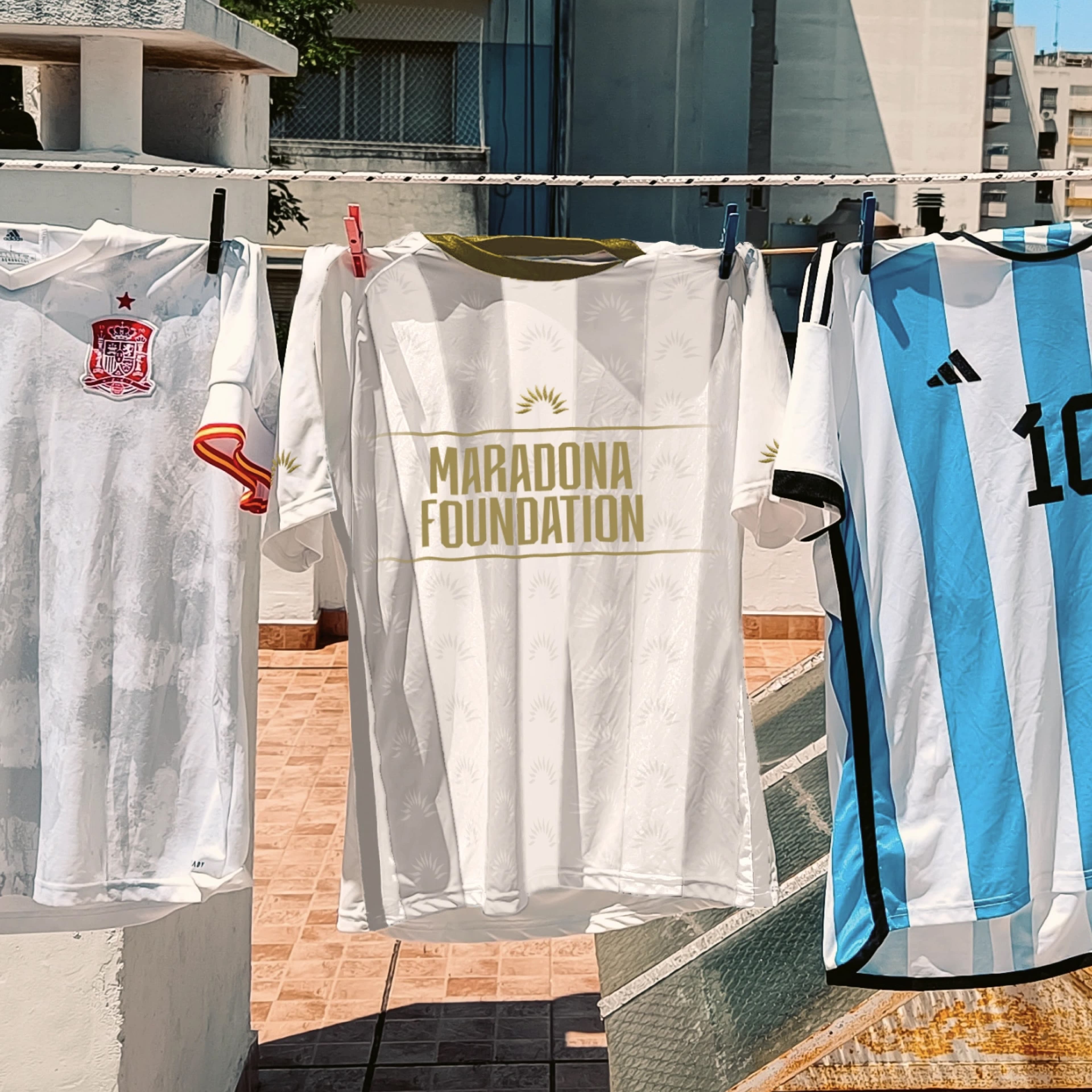 Three football jerseys hanging on a clothesline outdoors: a white Spain national team jersey with red crest, a white Maradona Foundation jersey with gold text and sunburst pattern, and a light blue and white striped Argentina Adidas jersey with number 10.