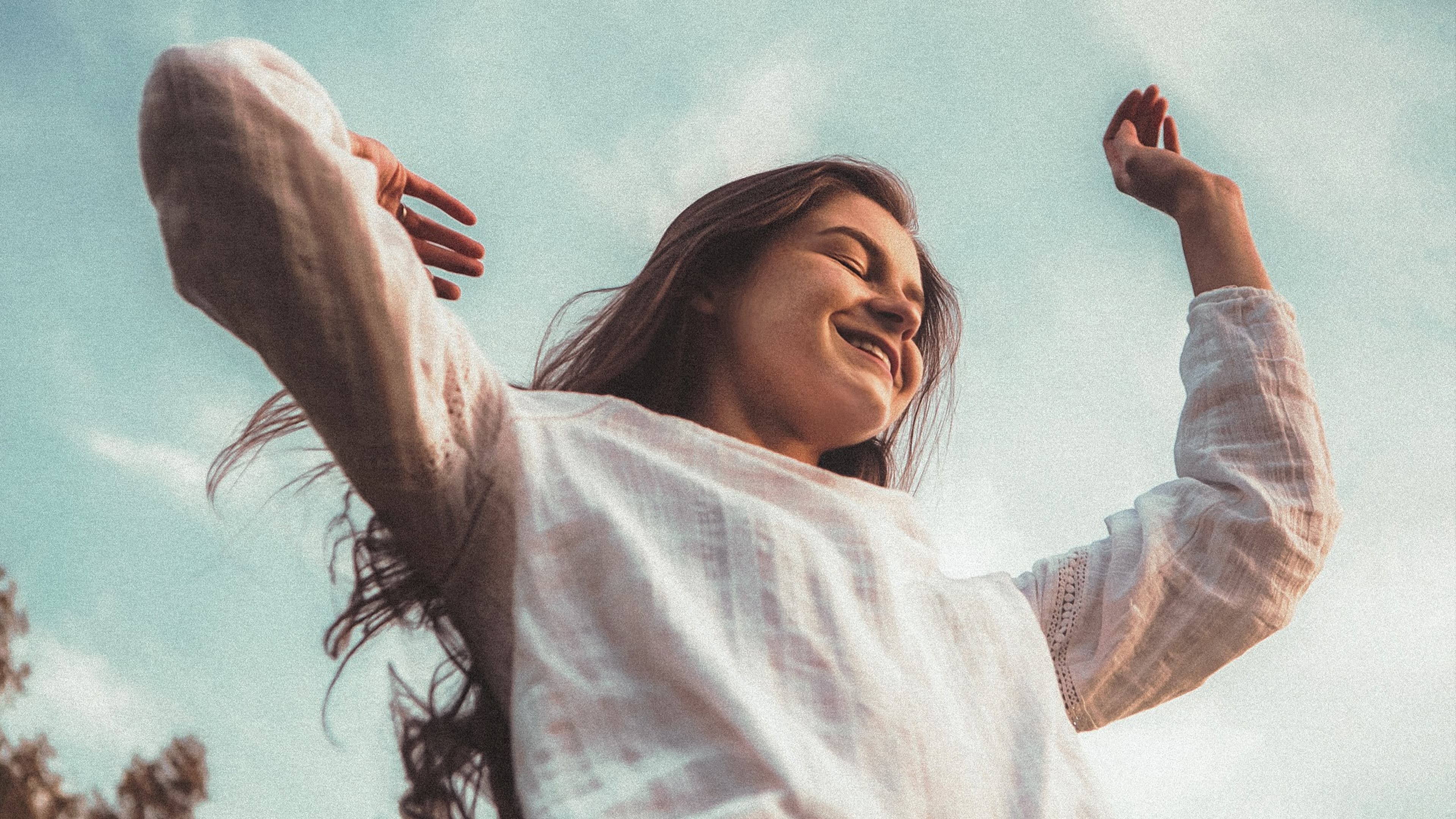 Young woman with long dark hair smiling joyfully with arms raised against a blue sky, wearing a white linen top