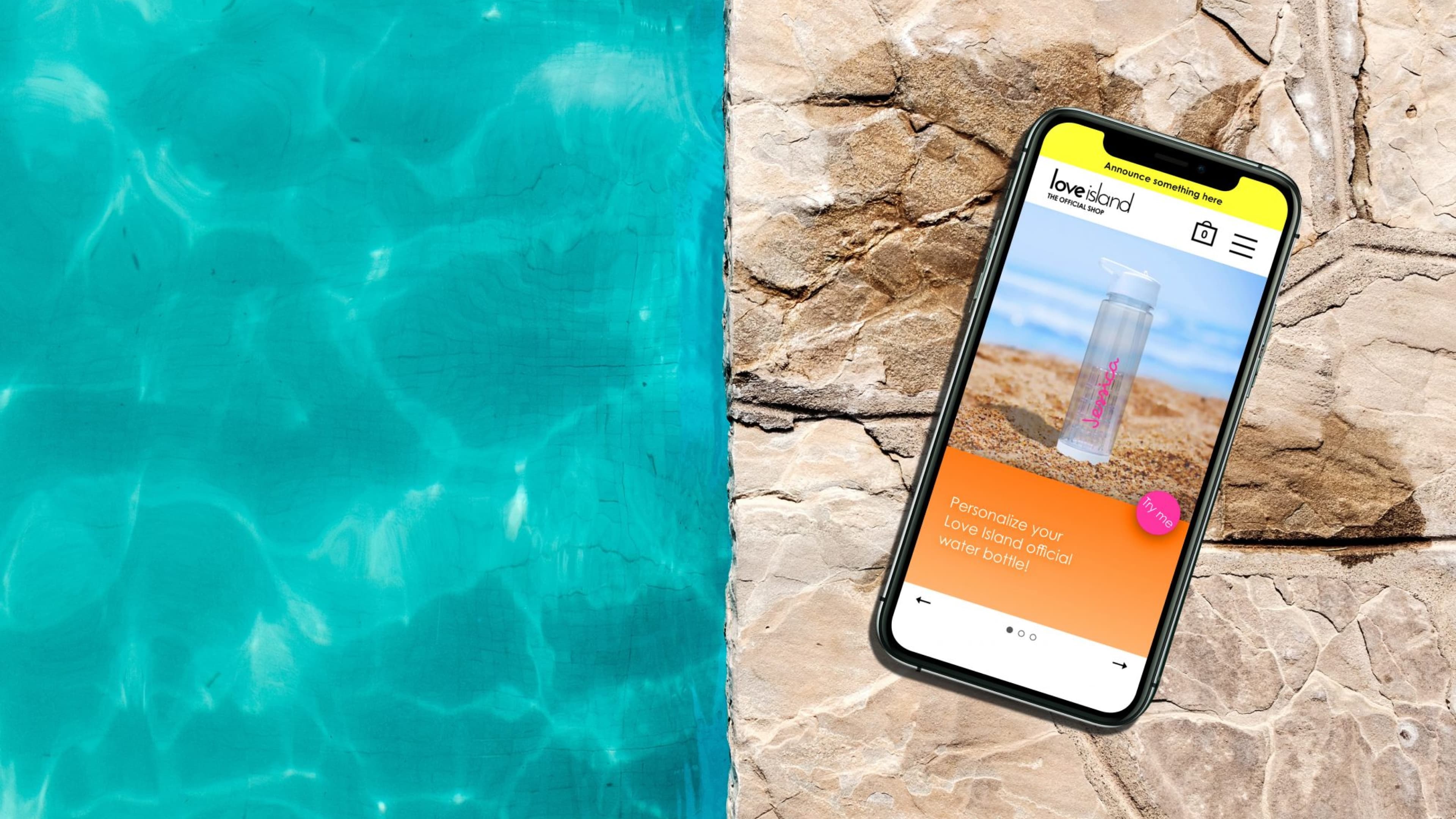 Smartphone displaying Love Island shop website with water bottle on rocky surface next to vibrant blue swimming pool.