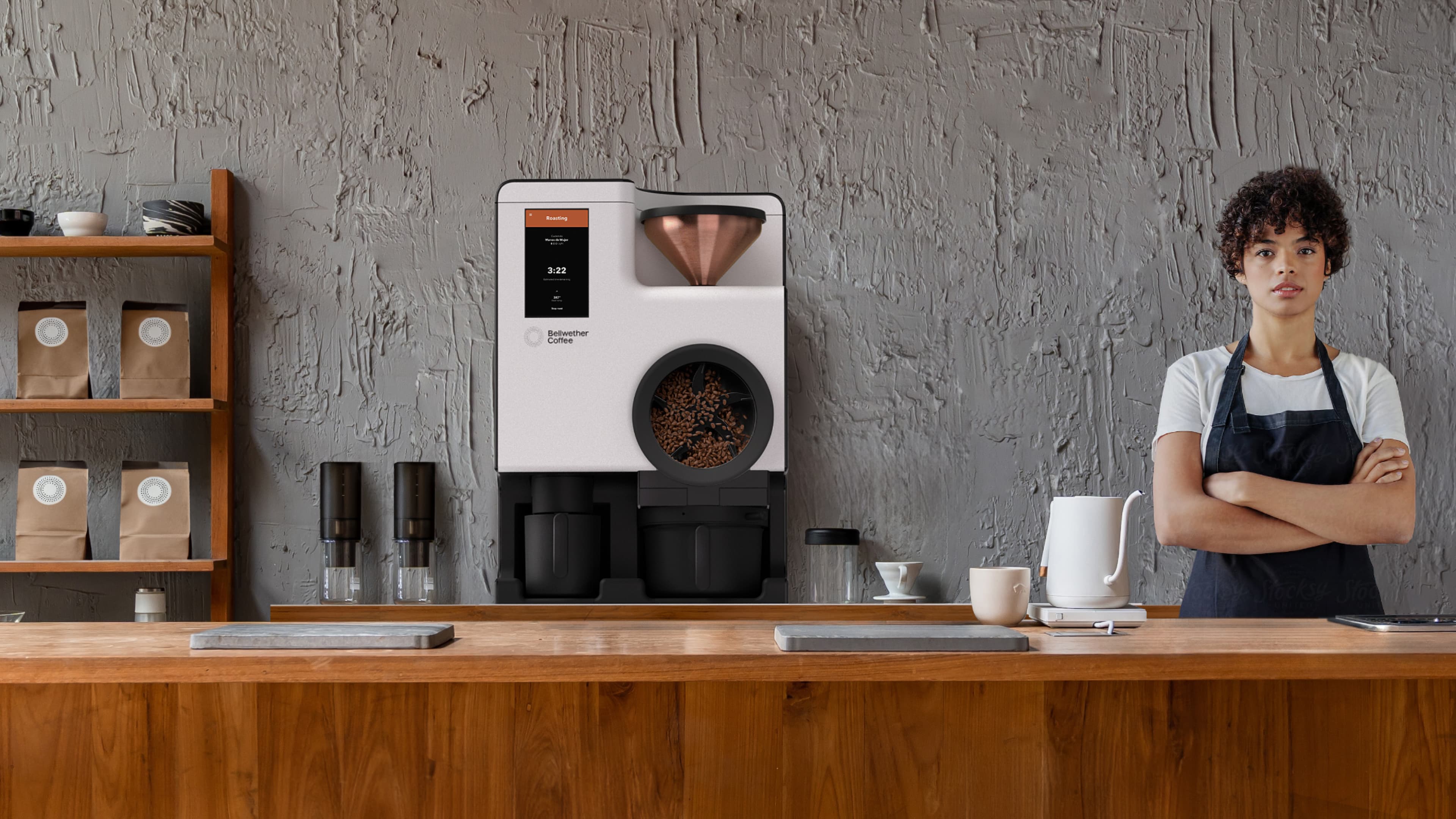 A Bellwether Coffee electric roaster on a wooden café counter, displaying roasting progress on its touchscreen, with a barista standing beside it and coffee bags on shelves behind.
