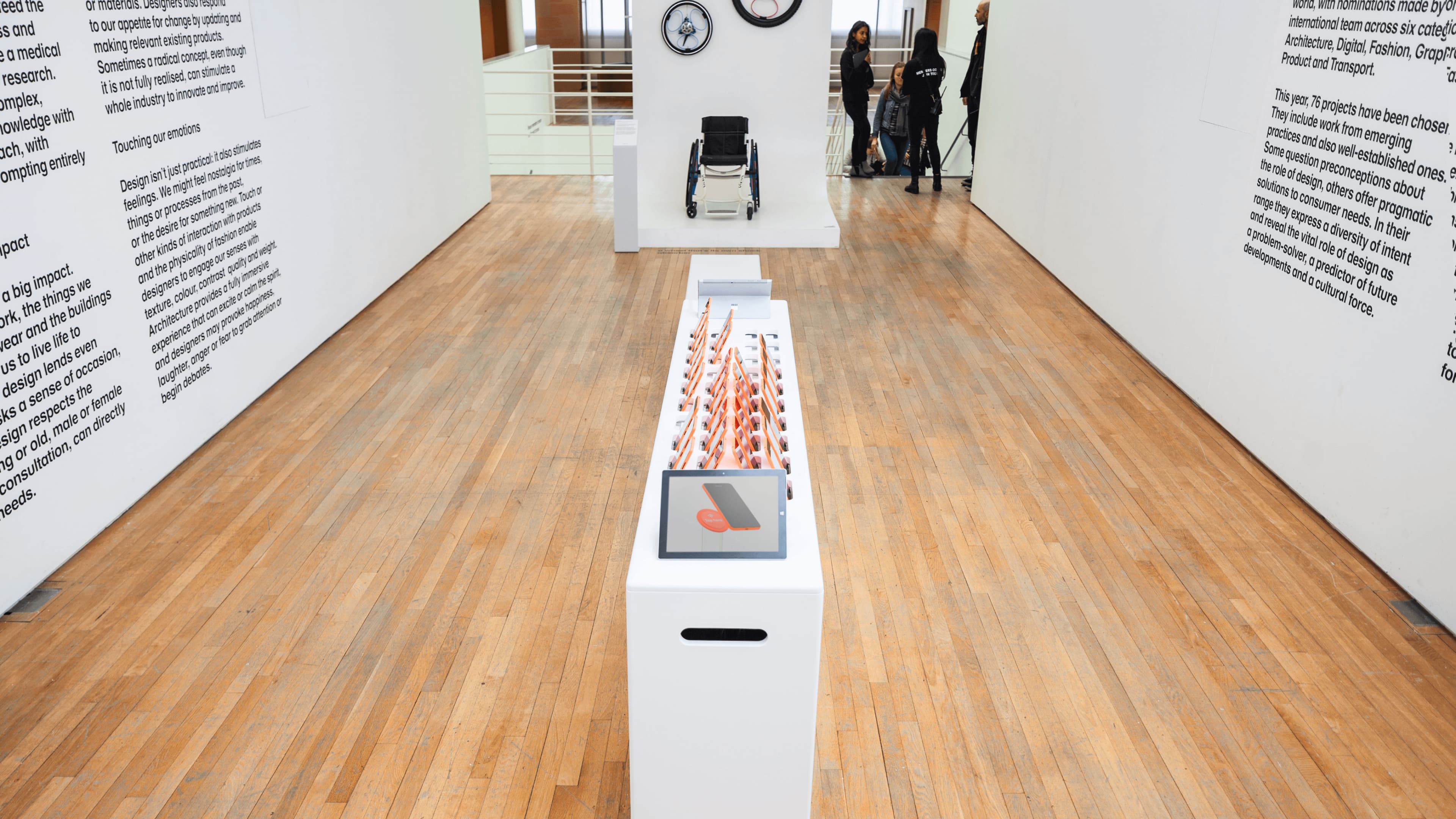 Interior of a design exhibition gallery showing a long white display case filled with rows of orange-handled tools and a tablet screen, with text panels on the walls, a wheelchair exhibit on a pedestal, clocks, and visitors in the background.