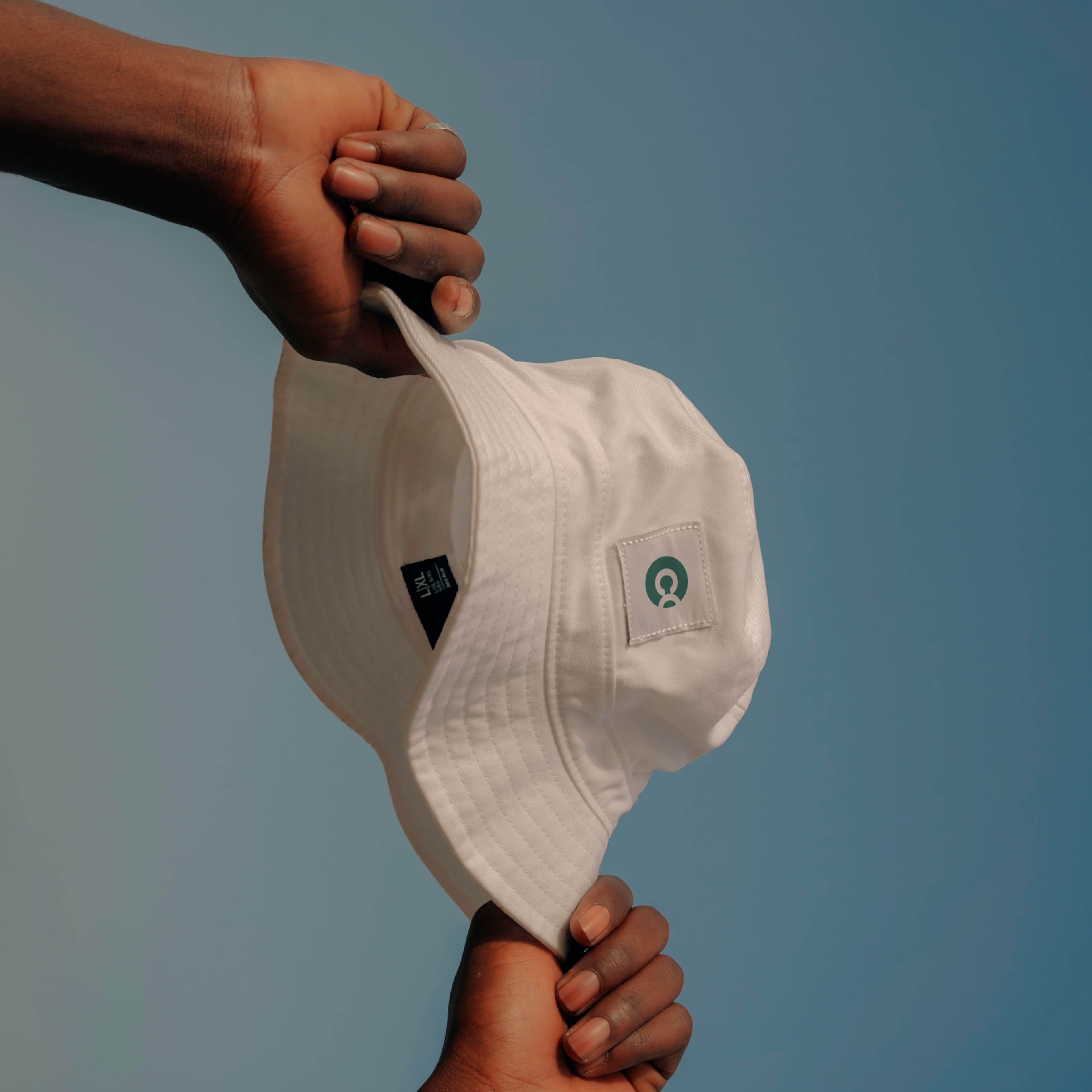 Two hands holding a white bucket hat against a clear blue sky. The hat displays a small green and white logo patch on the side.