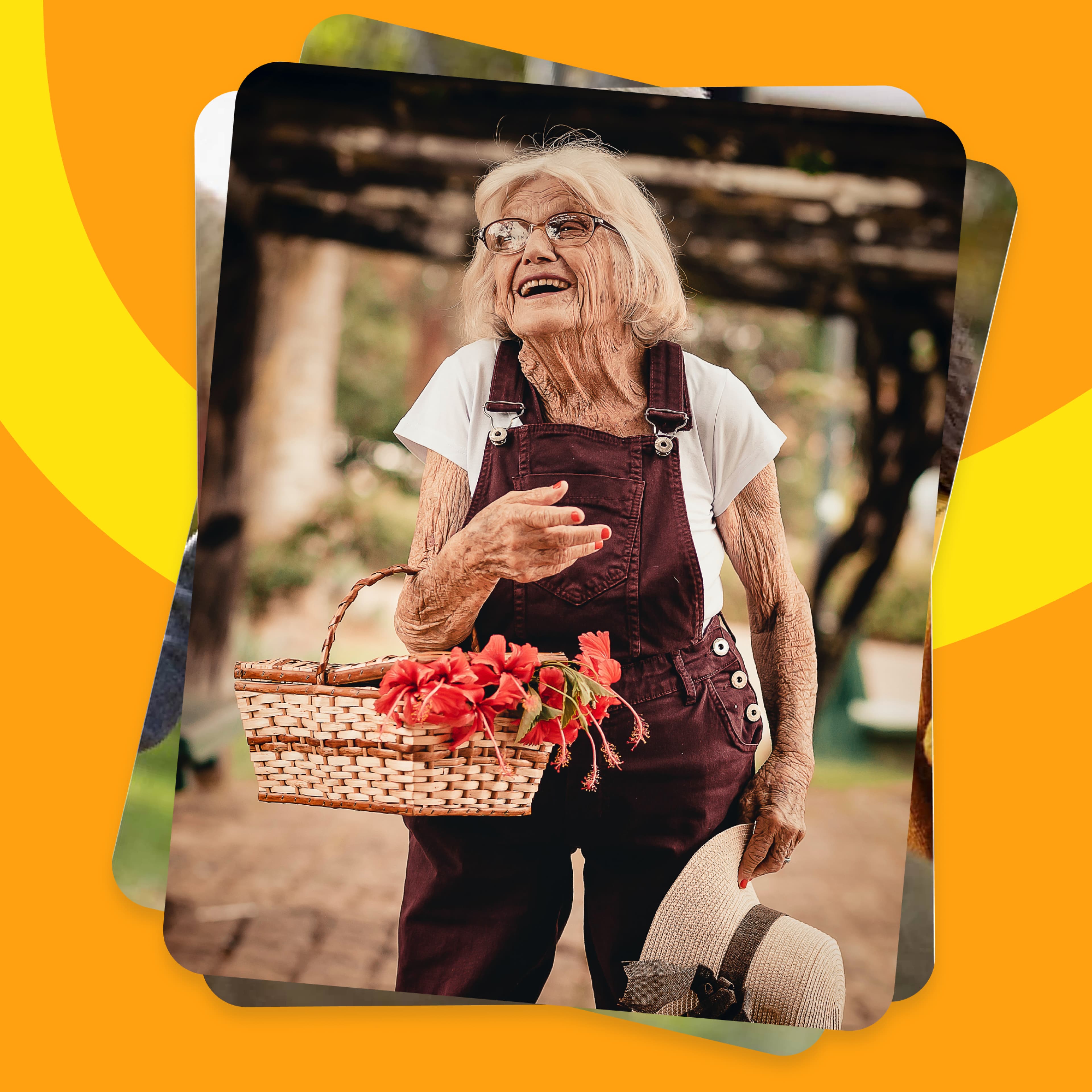 Cheerful elderly woman wearing glasses and maroon overalls, holding a wicker basket of red hibiscus flowers and a straw hat, laughing outdoors under a pergola in a garden.