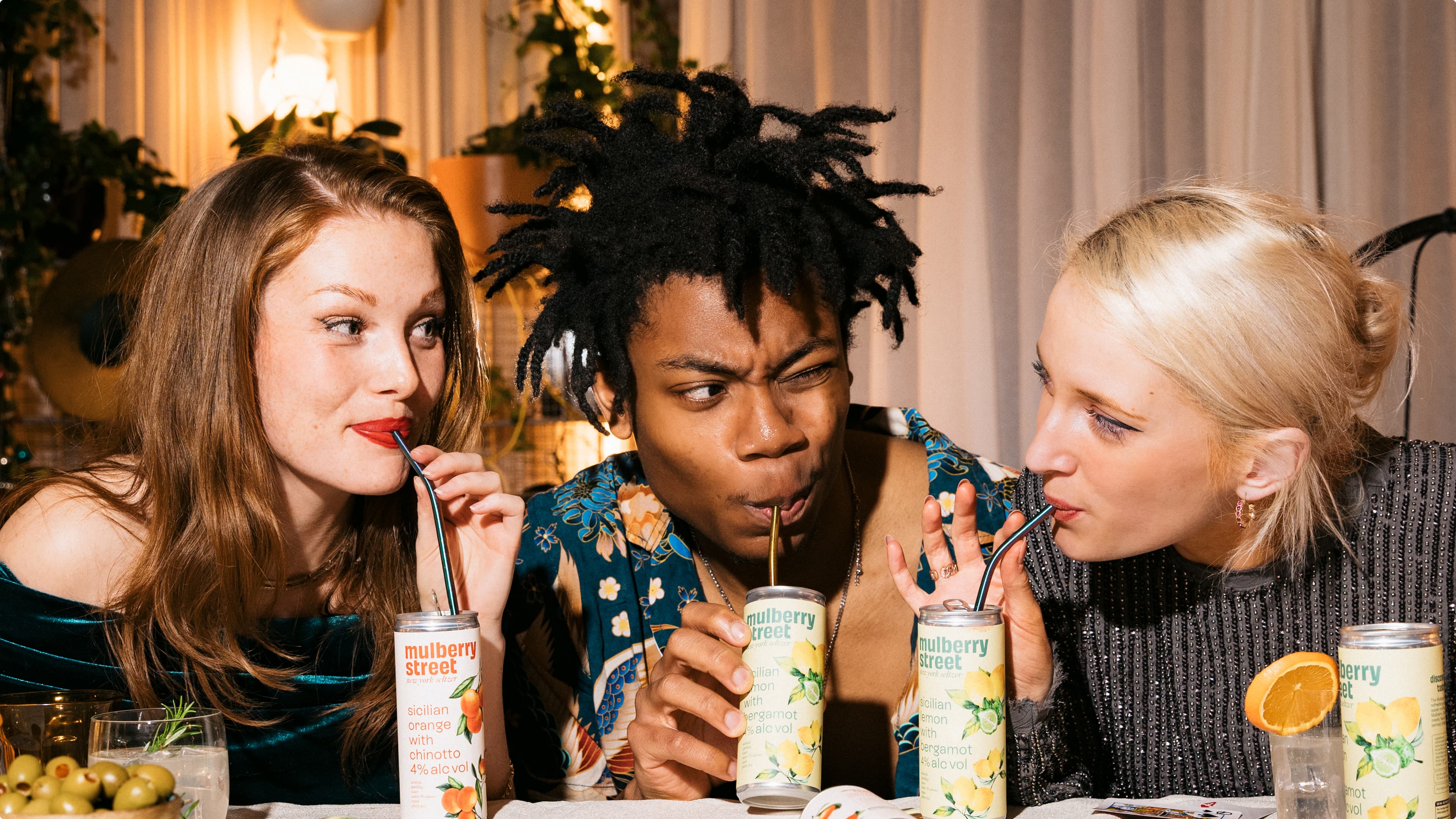Three people enjoying canned drinks with straws at a table. The drinks are from 'Mulberry Street'. Decor includes plants and soft lighting.