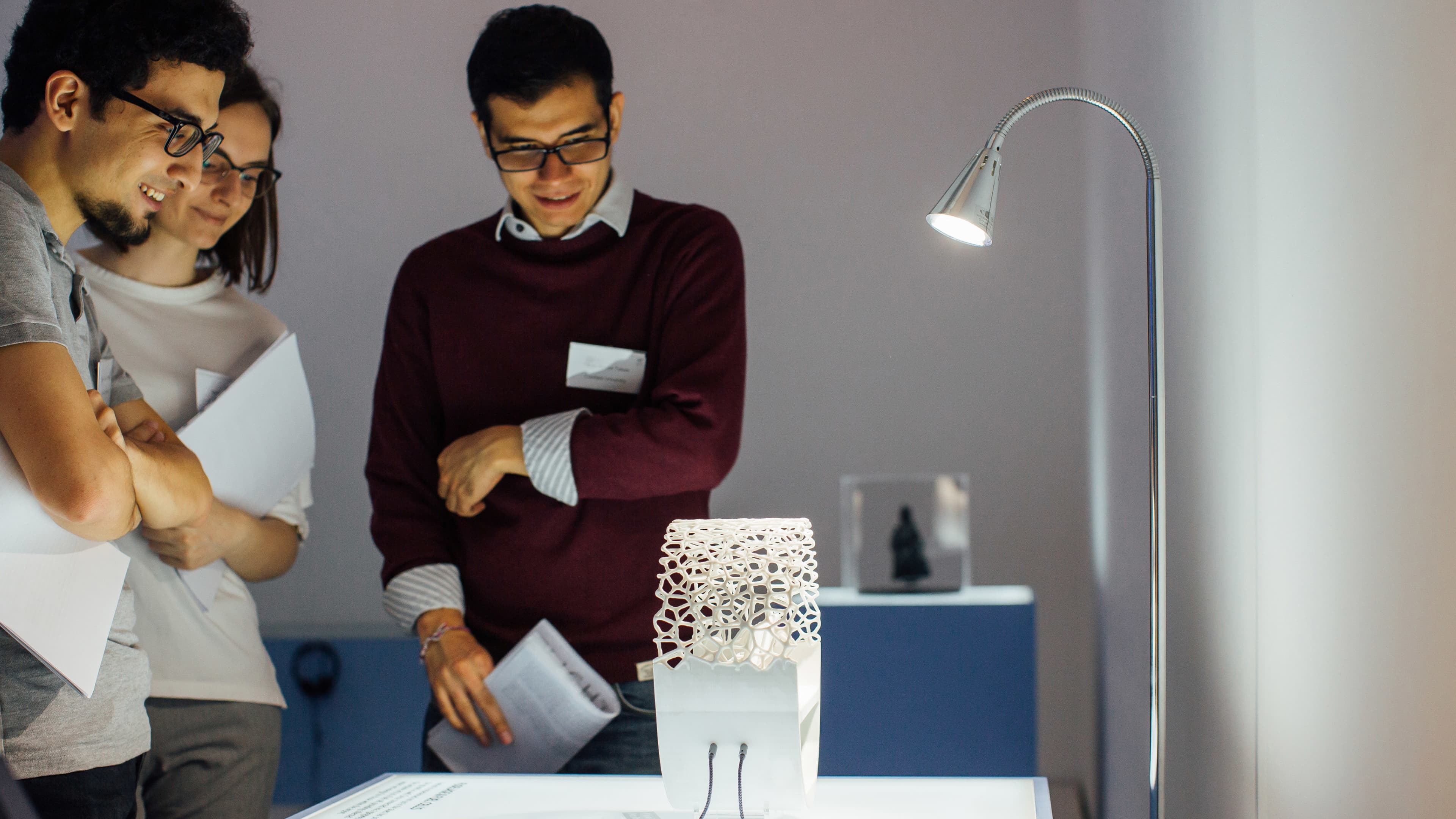 Three people smiling and examining a white 3D-printed architectural model on a lit display table in a design exhibition, holding papers.